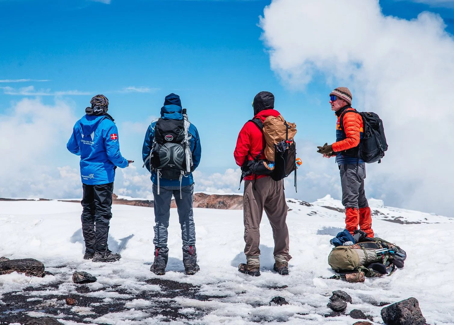 Four hikers dressed in winter gear, with backpacks, standing on snow-covered ground under a bright blue sky with clouds, appearing to be on a mountain or high-altitude location.