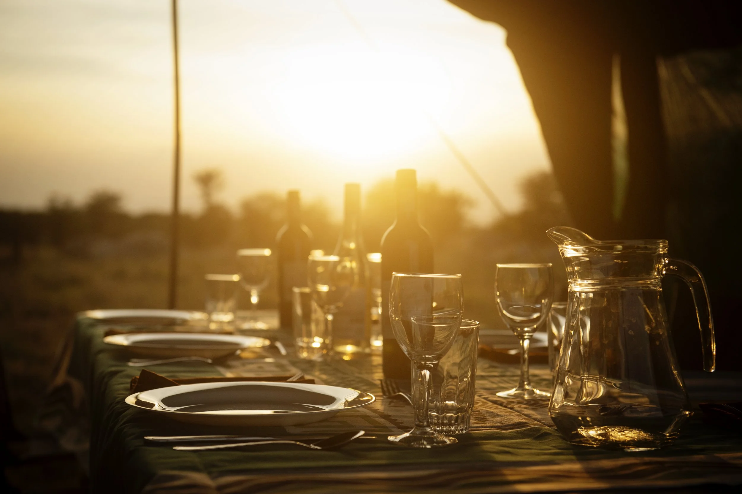 Dining table set with plates, glasses, and a pitcher, illuminated by warm sunlight, with a blurred outdoor background.