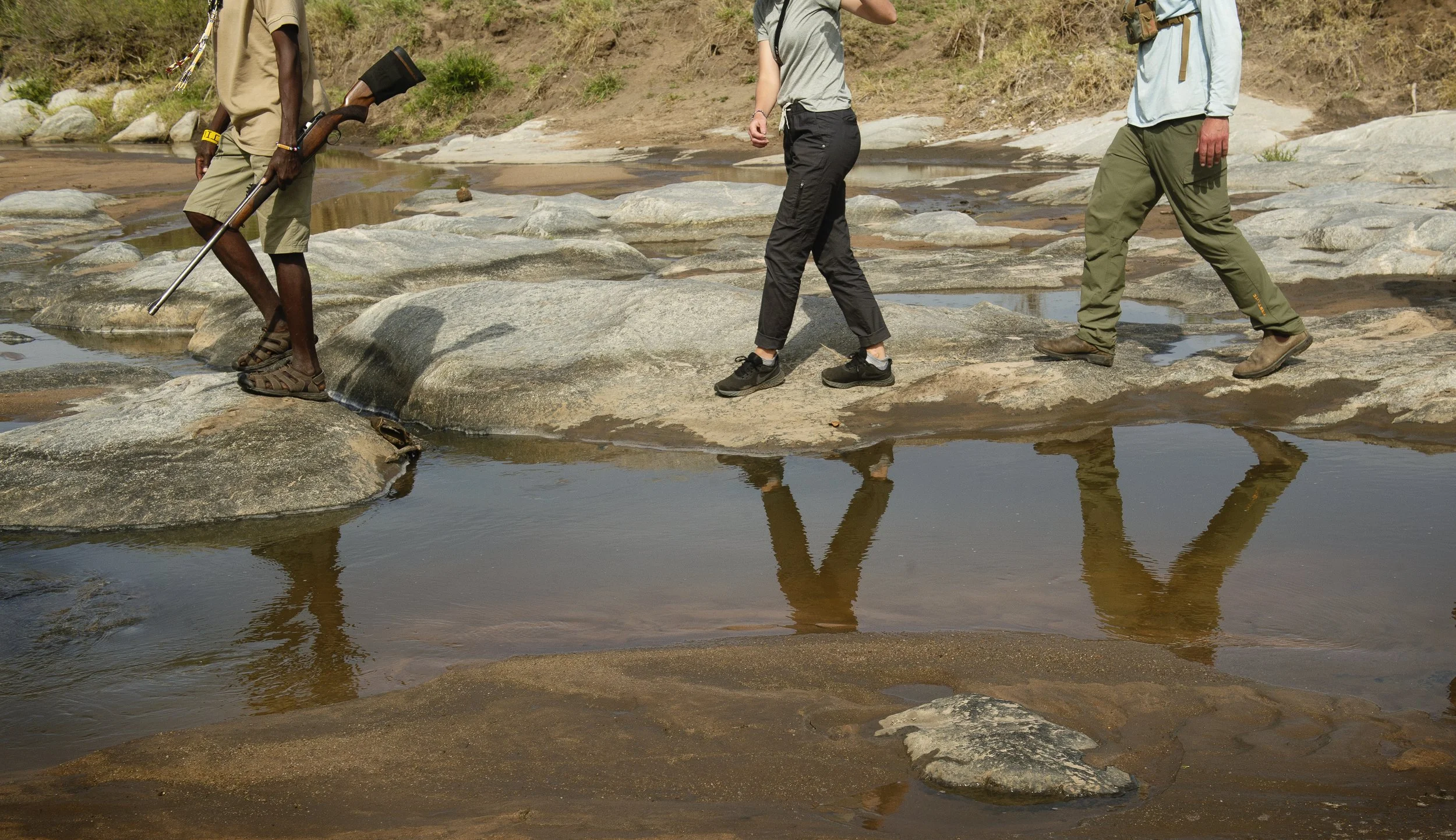 People crossing a shallow river over large rocks, with reflections visible in the water, outdoors on a sunny day.
