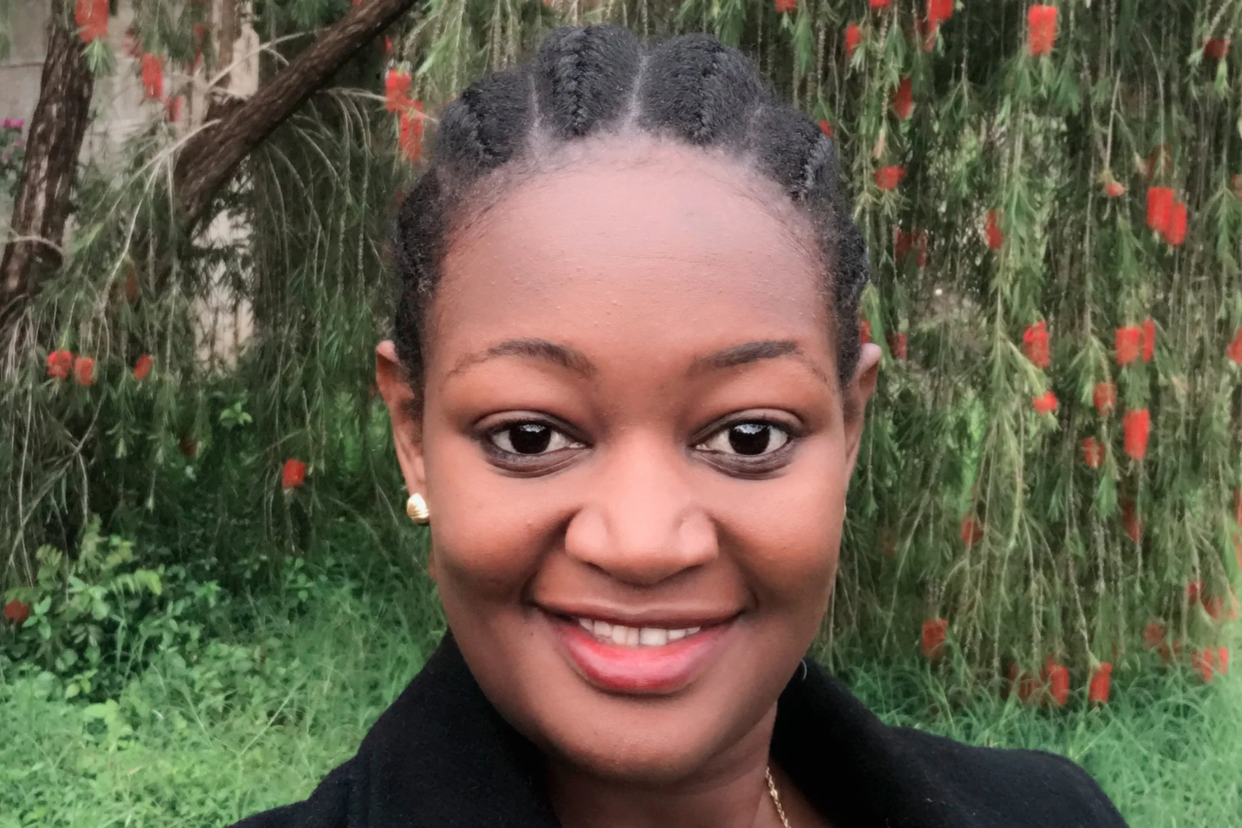 Close-up of a smiling woman with braided hair, wearing earrings and a dark top, outdoors with green grass and flowering plants in the background.