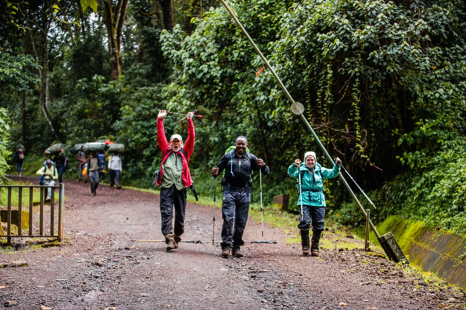 Group of hikers walking on a trail in a forest, one person holding a pole above the head in celebration, others smiling and carrying backpacks and hiking sticks.