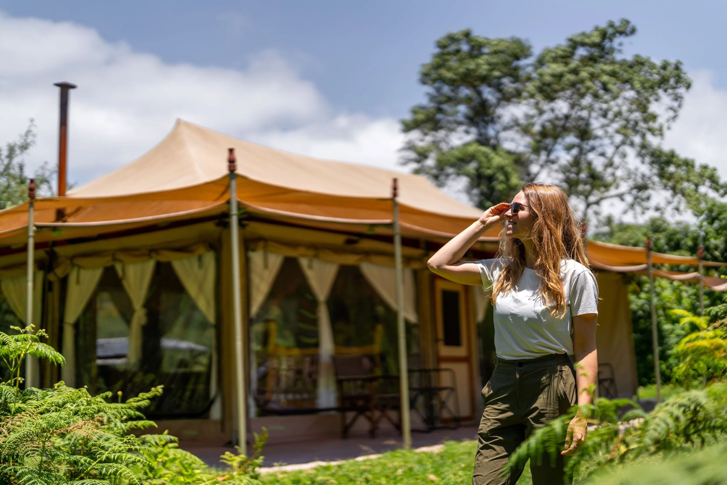 A young woman with long red hair wearing sunglasses, a white t-shirt, and olive-green pants, walking outdoors near a wooden pavilion with a beige canopy, surrounded by lush greenery and trees under a partly cloudy sky.