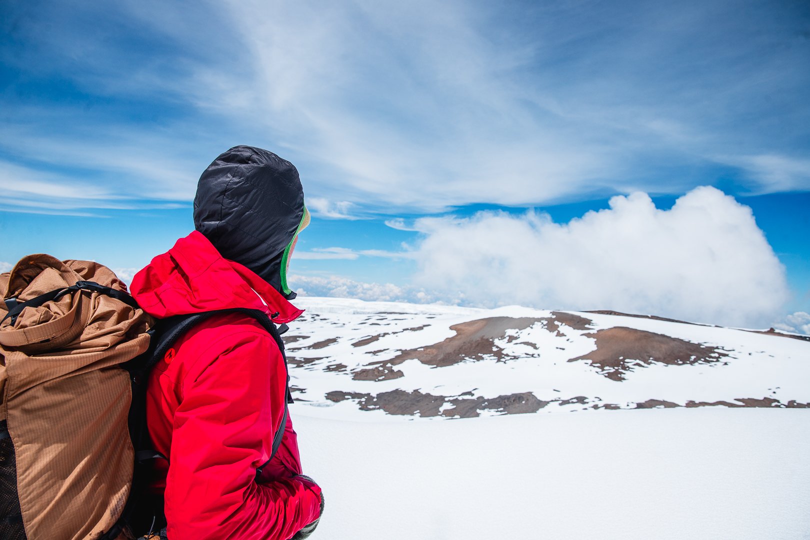 A person dressed in a red jacket and black hood, carrying a brown backpack, standing on a snowy mountain with a partly cloudy sky in the background.
