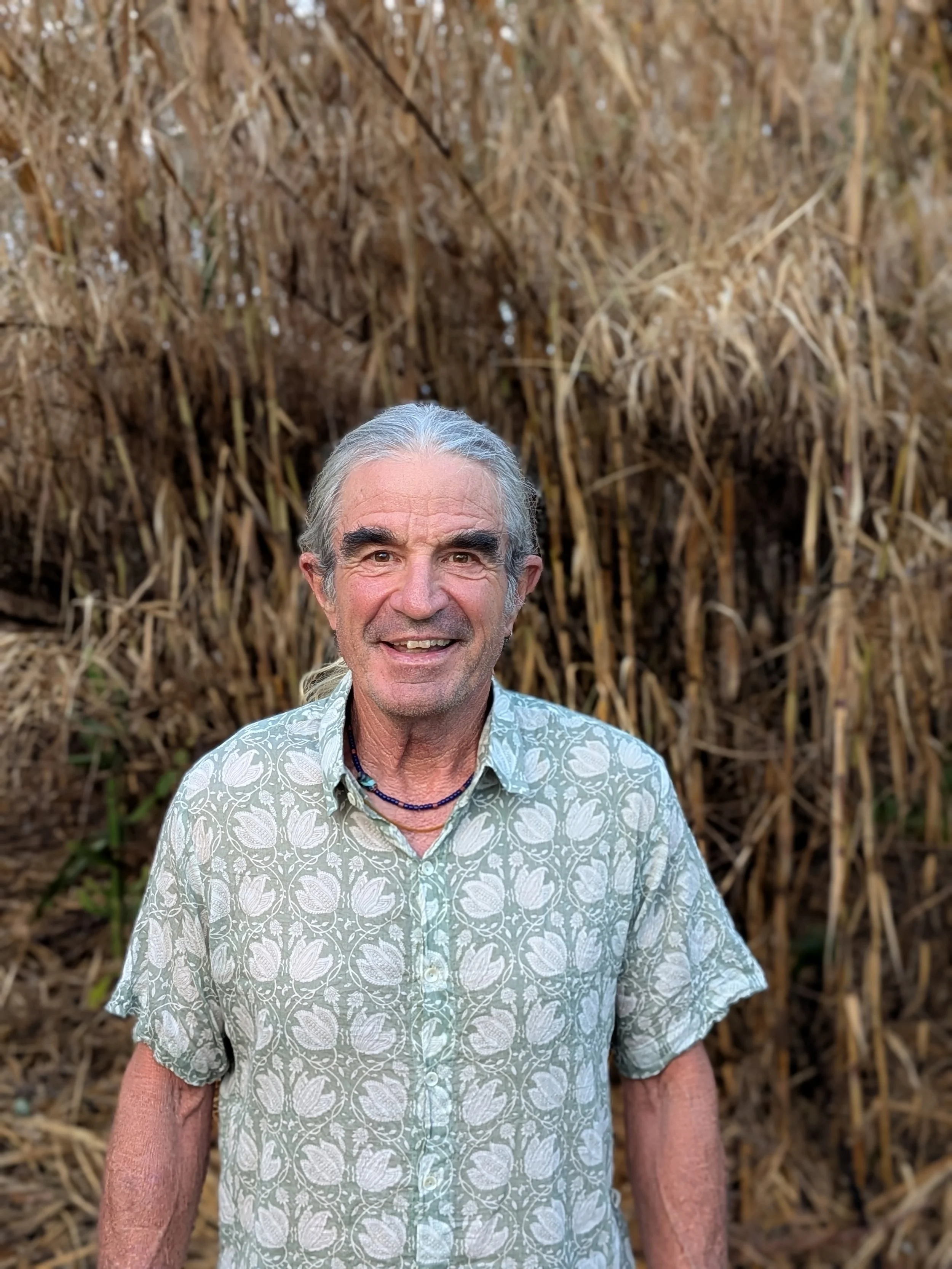 A middle-aged man with gray hair and a floral patterned short-sleeve shirt standing outdoors in front of a large pile of dried, brown plant stalks or hay, smiling at the camera.