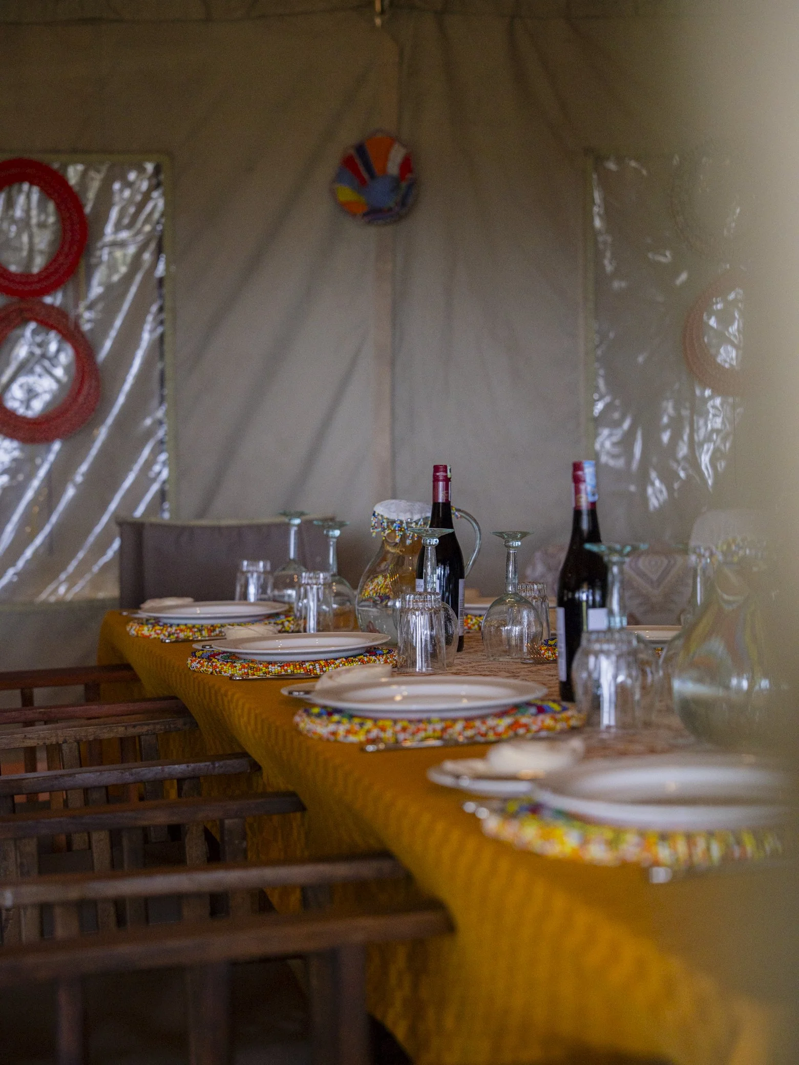 A dining table set with plates, glasses, bottles of wine, and colorful beaded placemats, with a wall decorated with hanging hoops and a colorful round art piece.