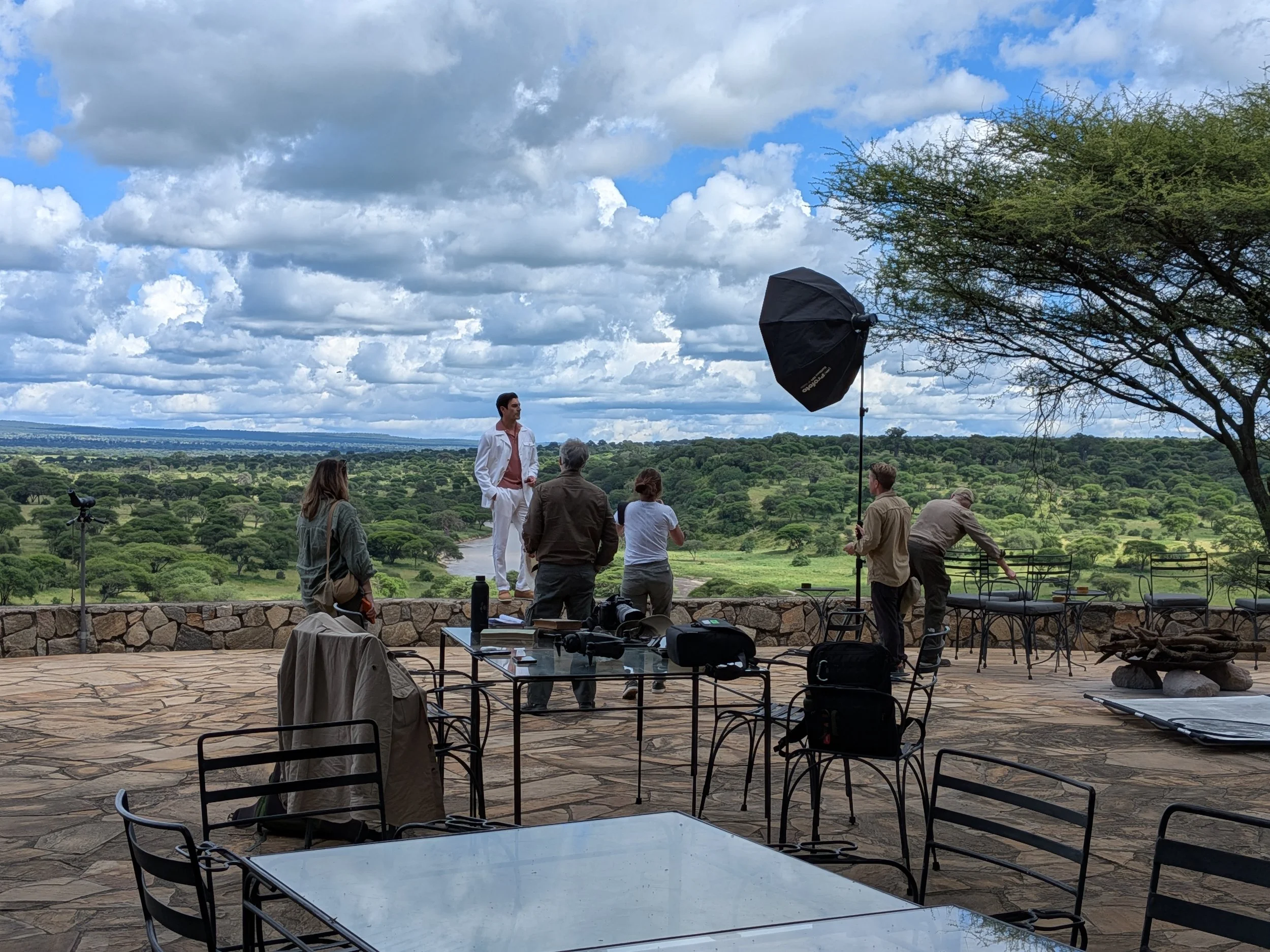 A photoshoot setup outdoors on a stone patio overlooking a landscape of green trees under a partly cloudy sky. There is a person in white clothing standing near a group of seated people, with professional lighting equipment visible.
