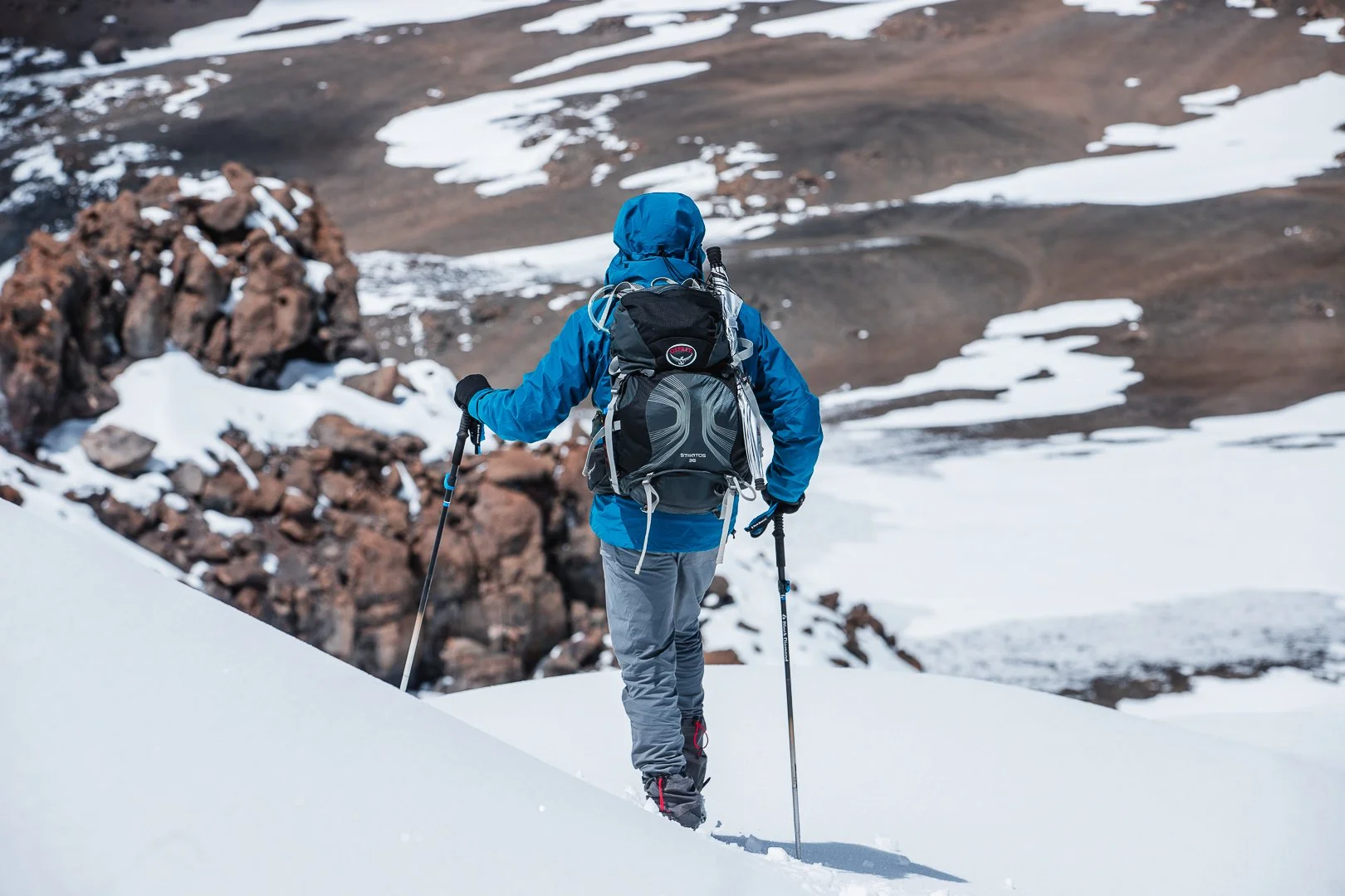 A hiker in a blue jacket and gray pants trekking through snow with hiking poles in a snowy mountainous landscape.