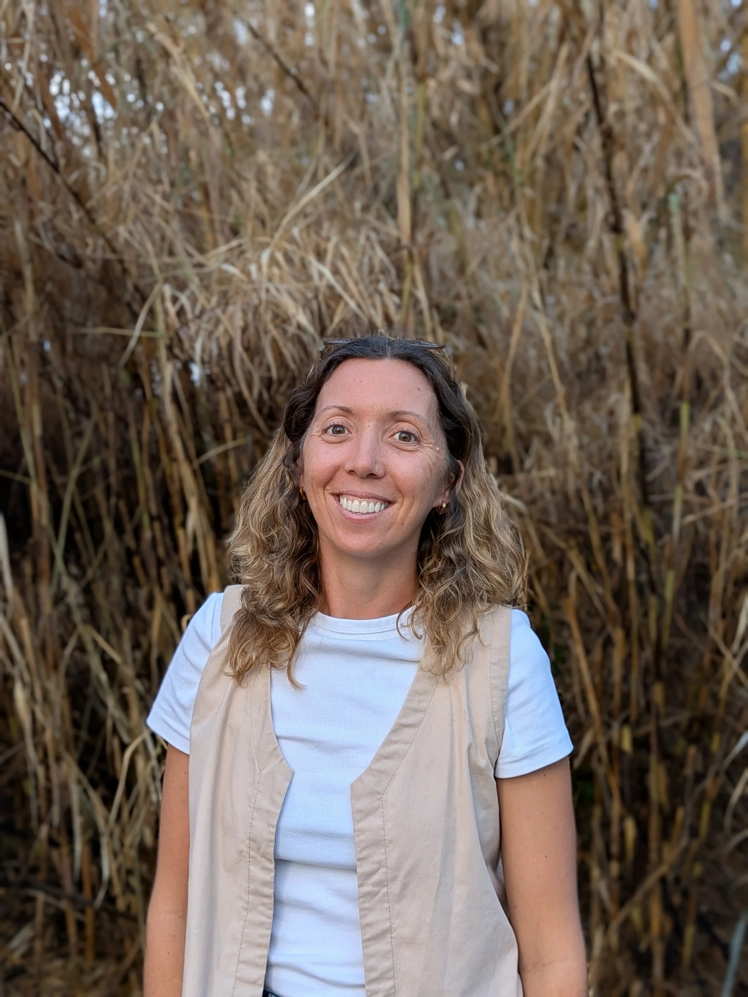 A woman with long, curly hair smiling at camera, standing outdoors in front of dried corn stalks.