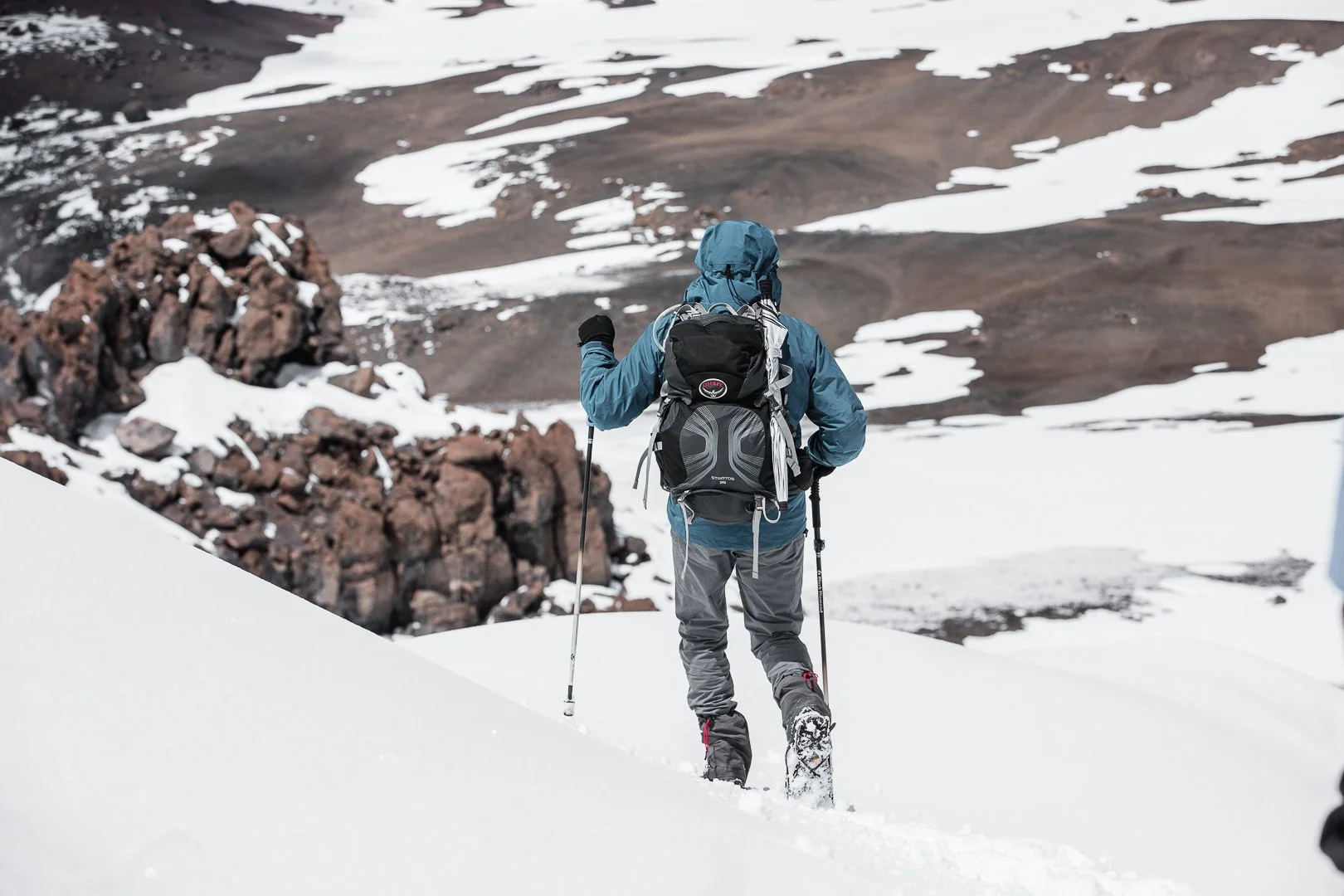 A person hiking through snow-covered terrain with rocky mountains and patches of snow in the background, wearing a blue jacket, black backpack, and using trekking poles.