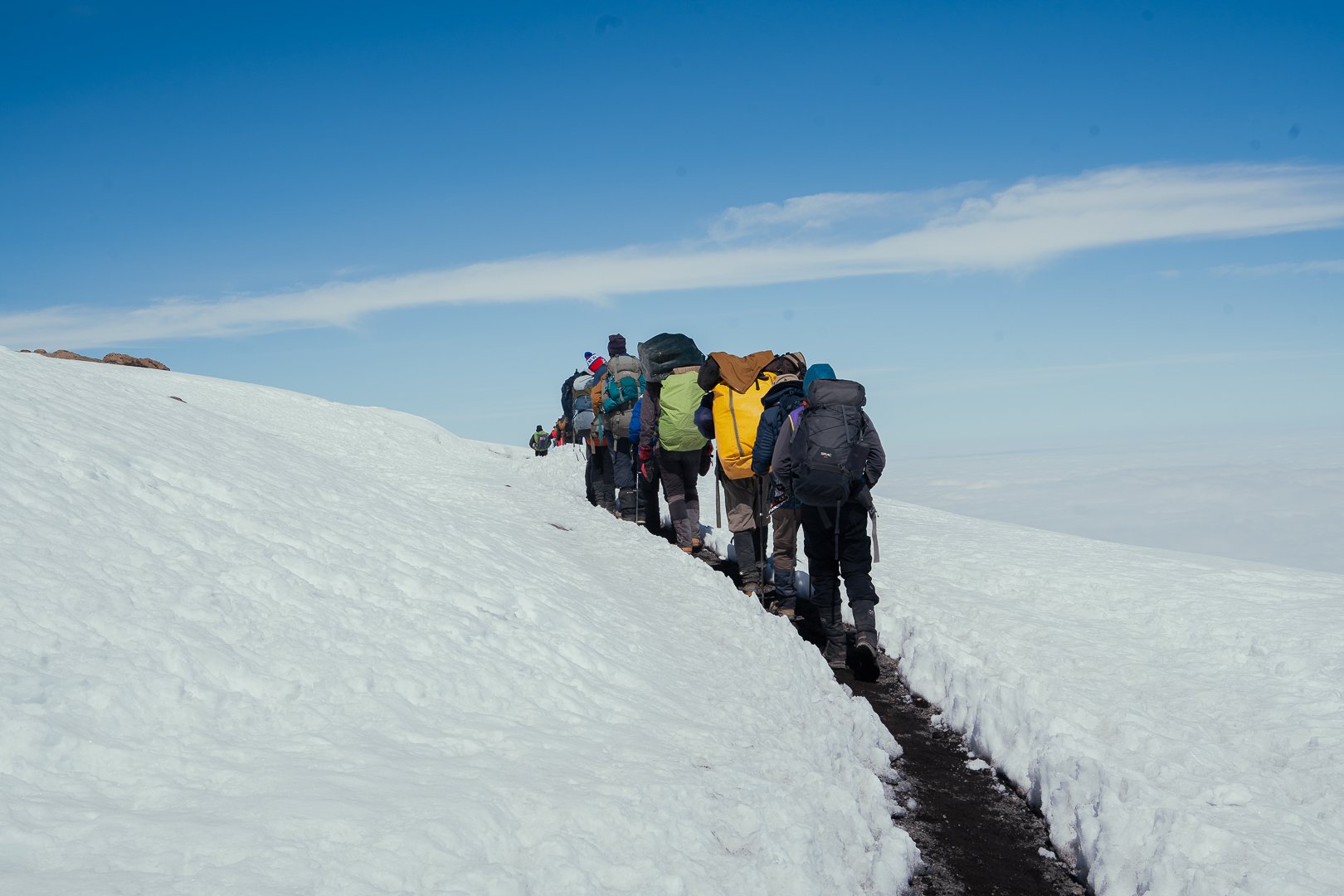 A group of hikers in winter gear walking single file on a snow-covered mountain trail with backpacks under a blue sky.