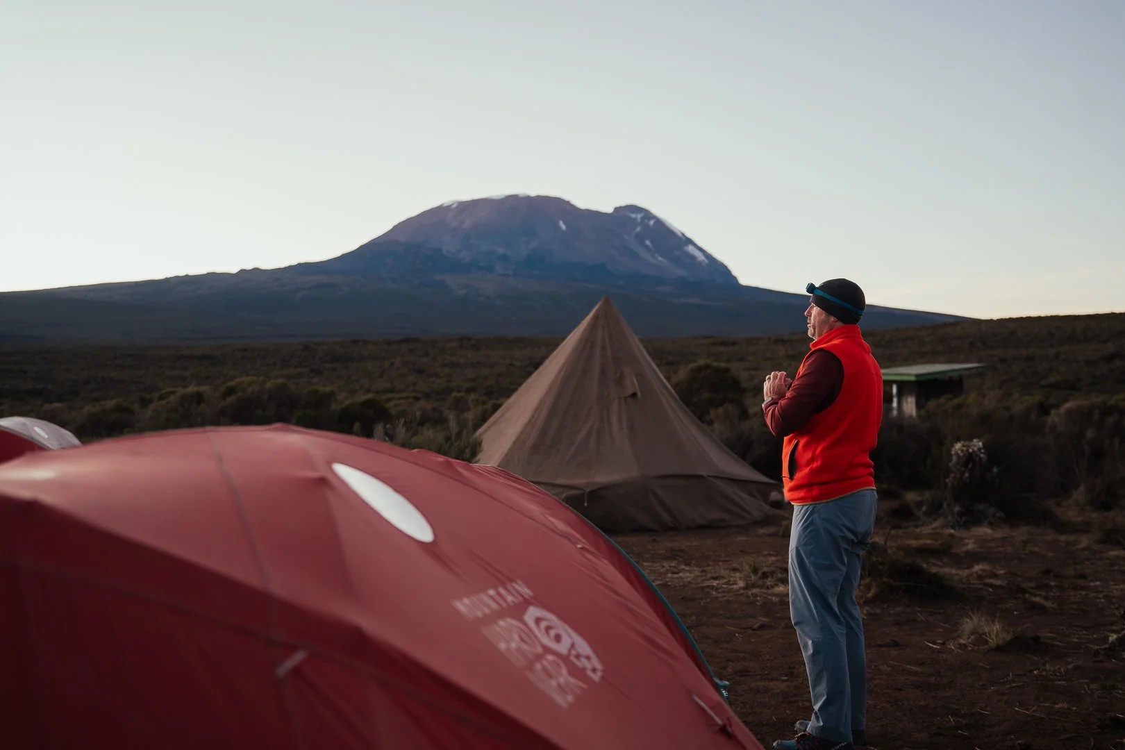 A man in outdoor gear, including a red vest and headlamp, stands with hands clasped looking at a mountain in the distance during sunset. Camping tents are set up in the foreground on a barren landscape.