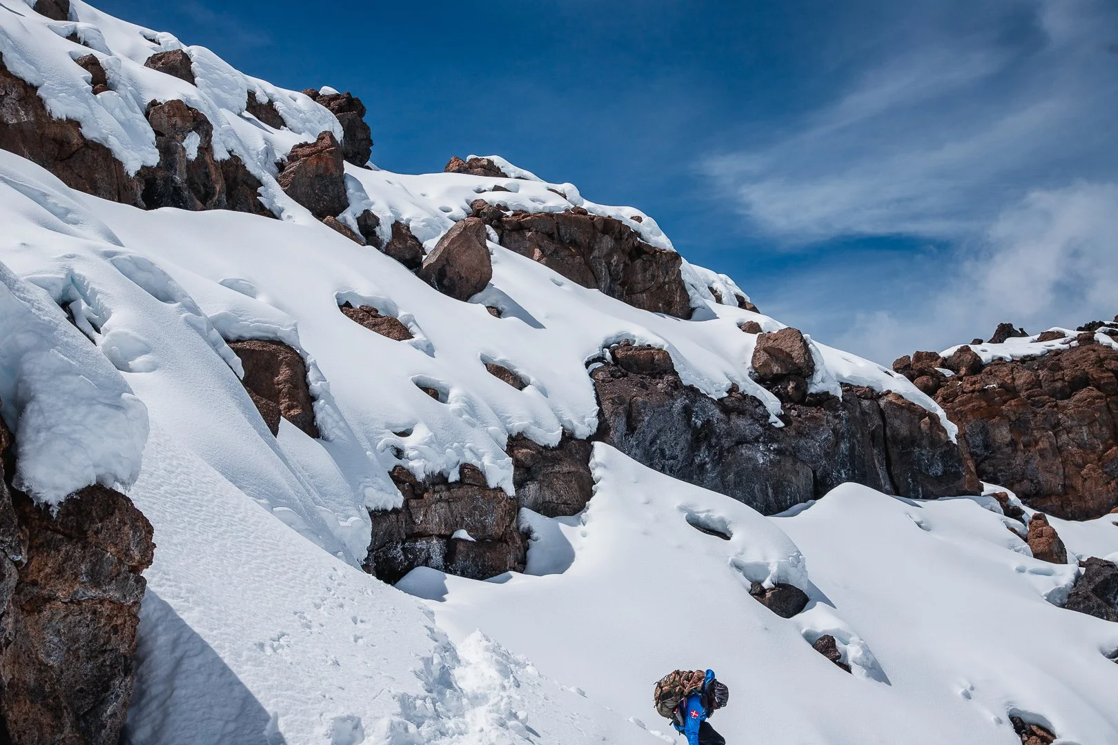 A person with a backpack hiking on a snow-covered mountain slope with large rocks and patches of snow, under a blue sky with some clouds.