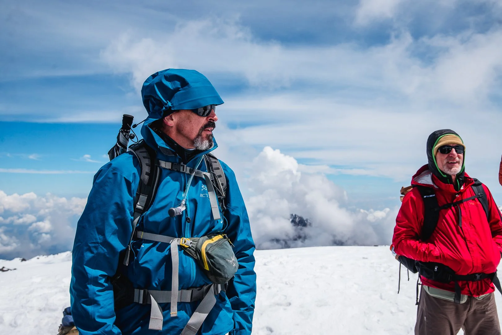 Two men in winter gear standing on snowy mountain landscape with clouds and blue sky in the background.