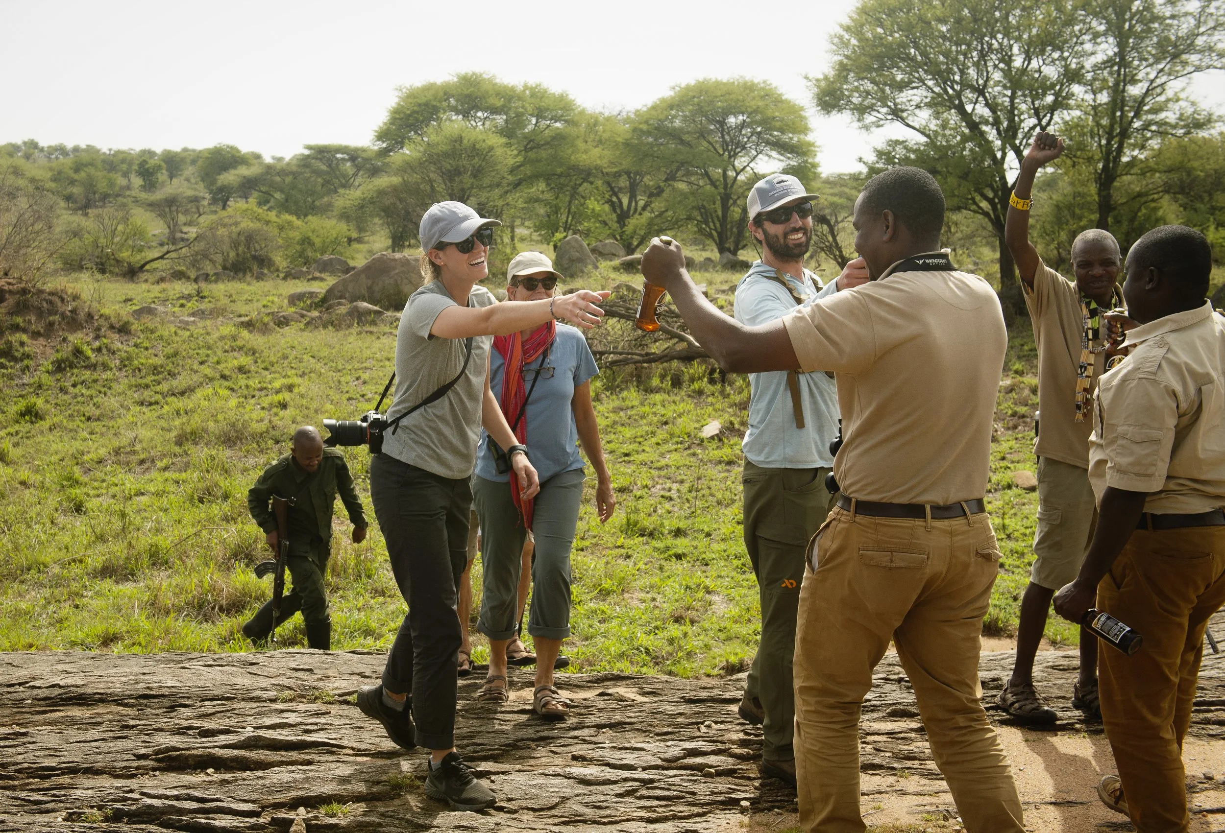 Group of tourists on safari in a savannah, some raising fists and others smiling, surrounded by green trees and rocks.
