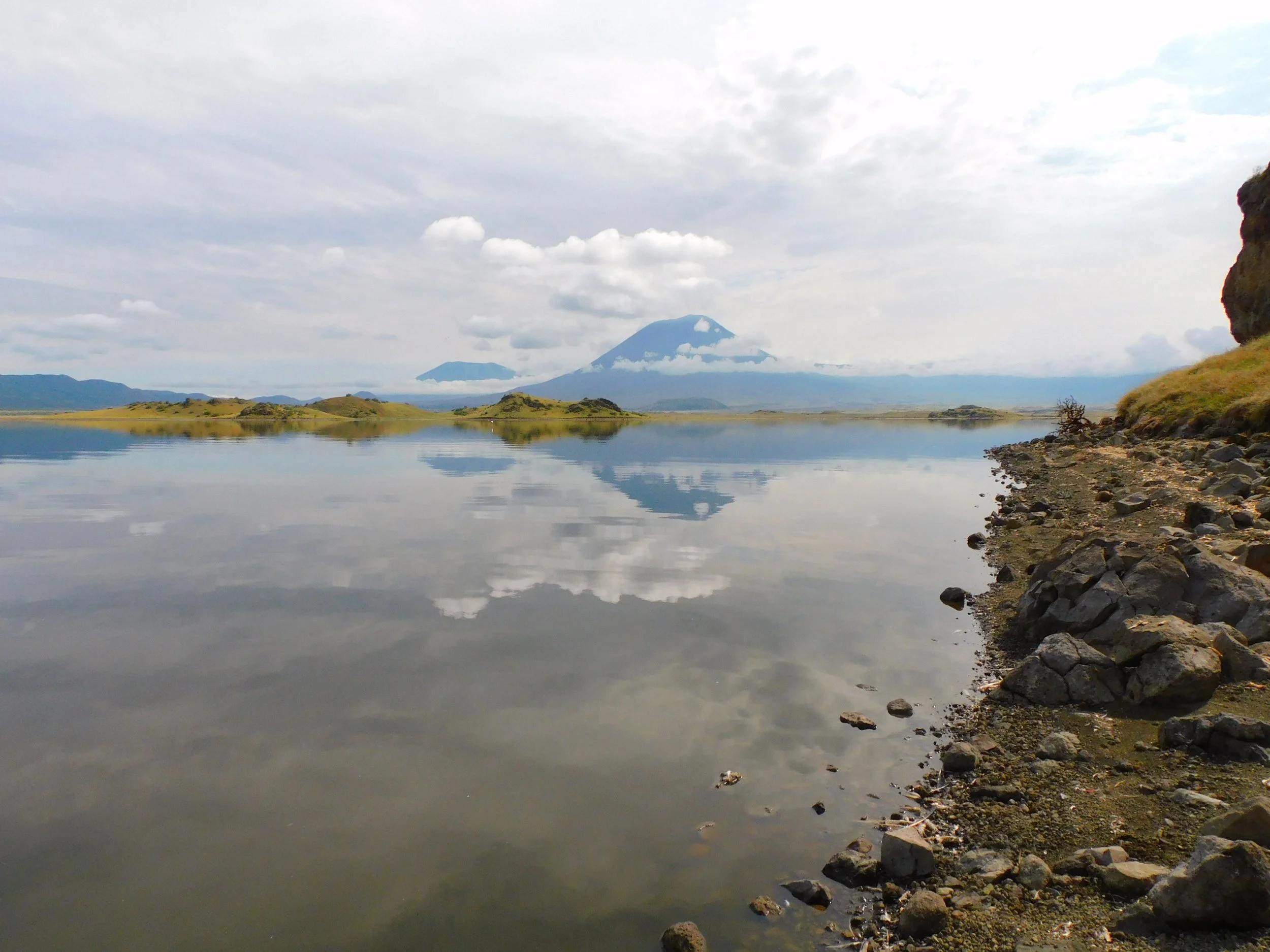 A calm lake reflecting the cloudy sky, with a mountain in the distance and a rocky shoreline on the right side of the image.