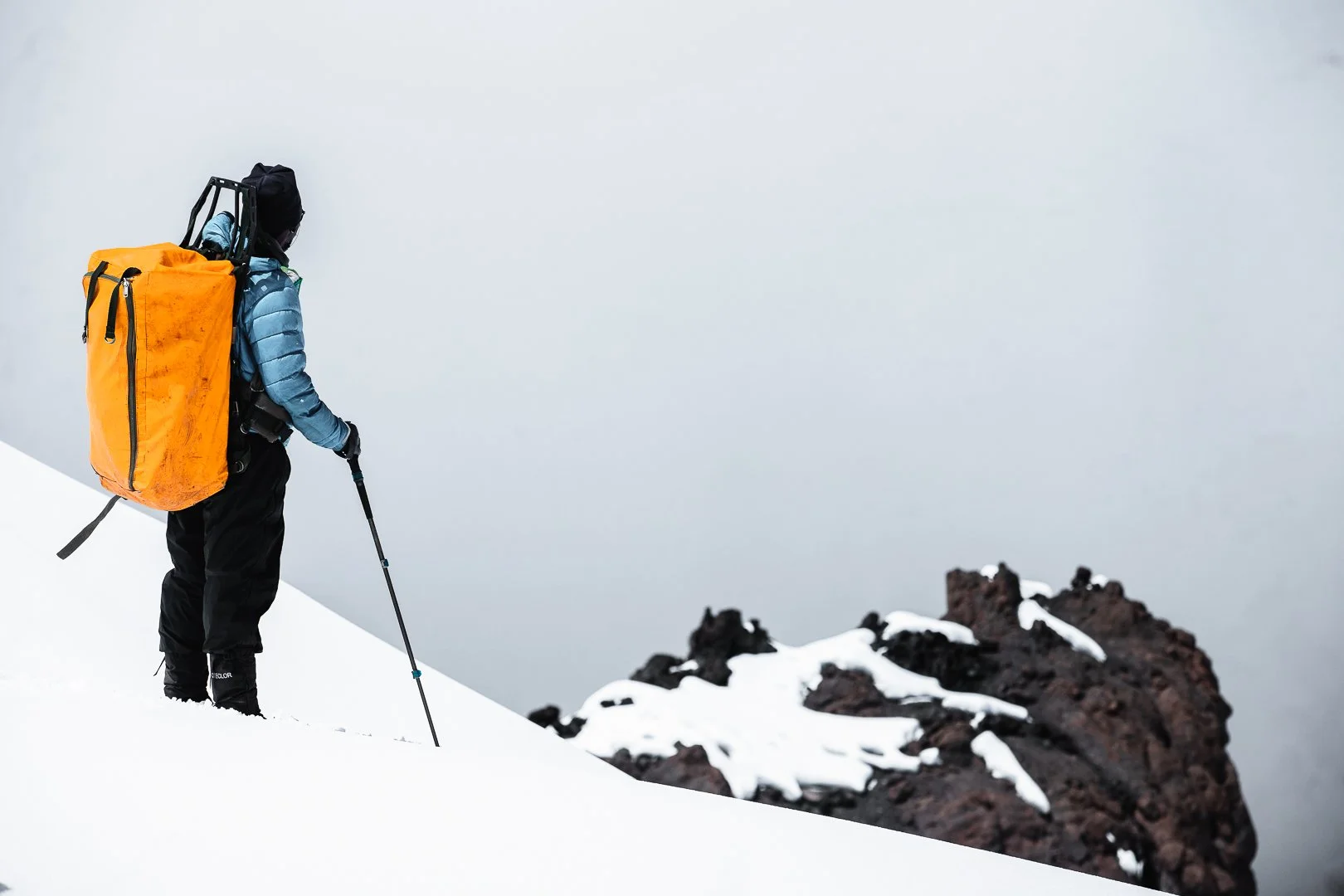 A person dressed in winter clothing with a yellow backpack, hiking on snow-covered terrain with black volcanic rocks in the background.