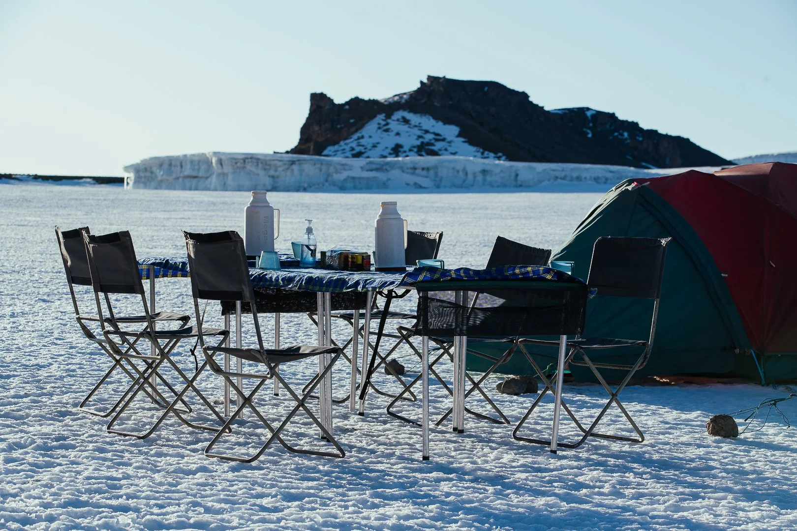 Camping setup with a table, chairs, water bottles, and a tent on snowy terrain with mountain-background.