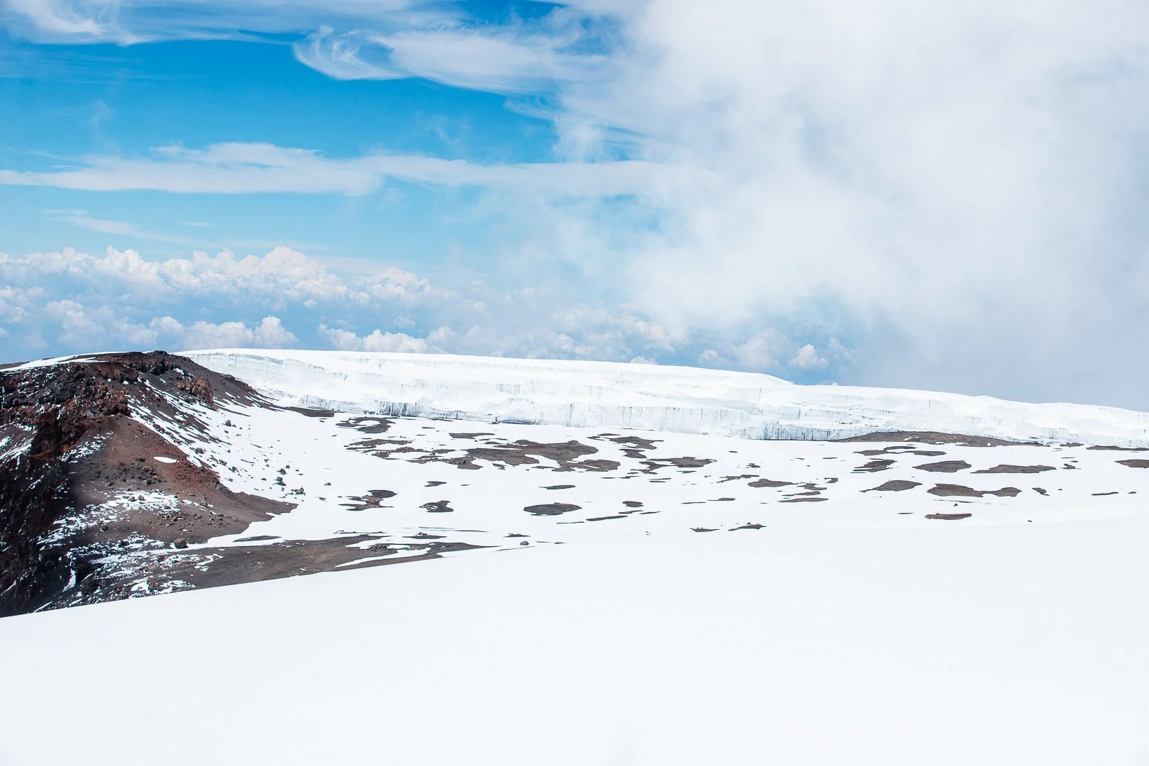 Snow-covered mountain landscape with a dark rocky area, patches of exposed earth, and a glacier under a blue sky with clouds.