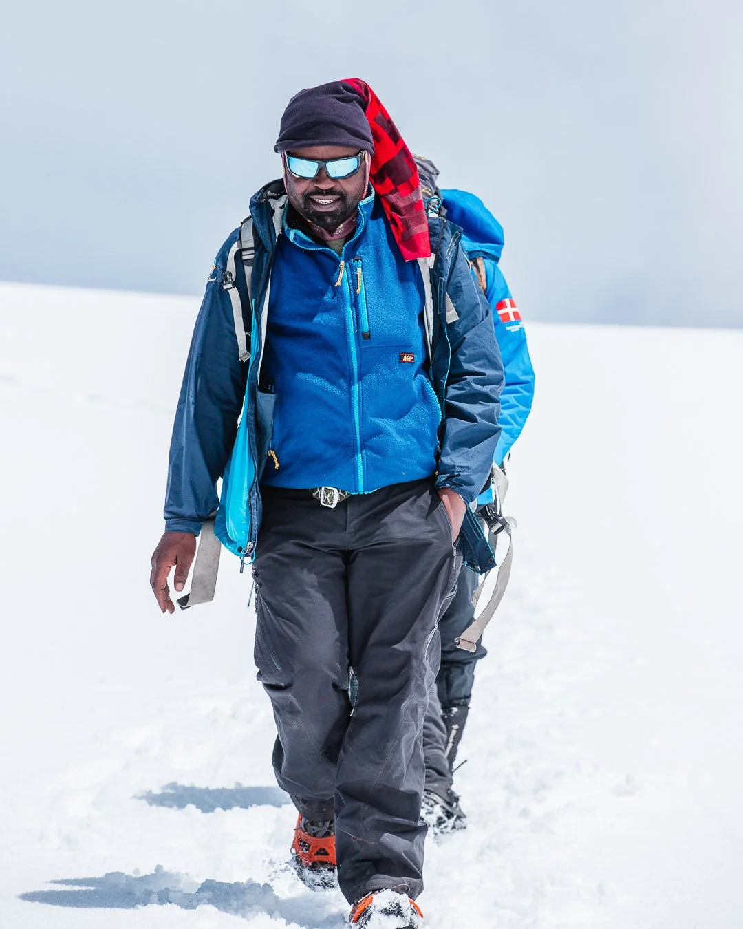 Two hikers walking in snow, wearing outdoor gear, sunglasses, and backpacks