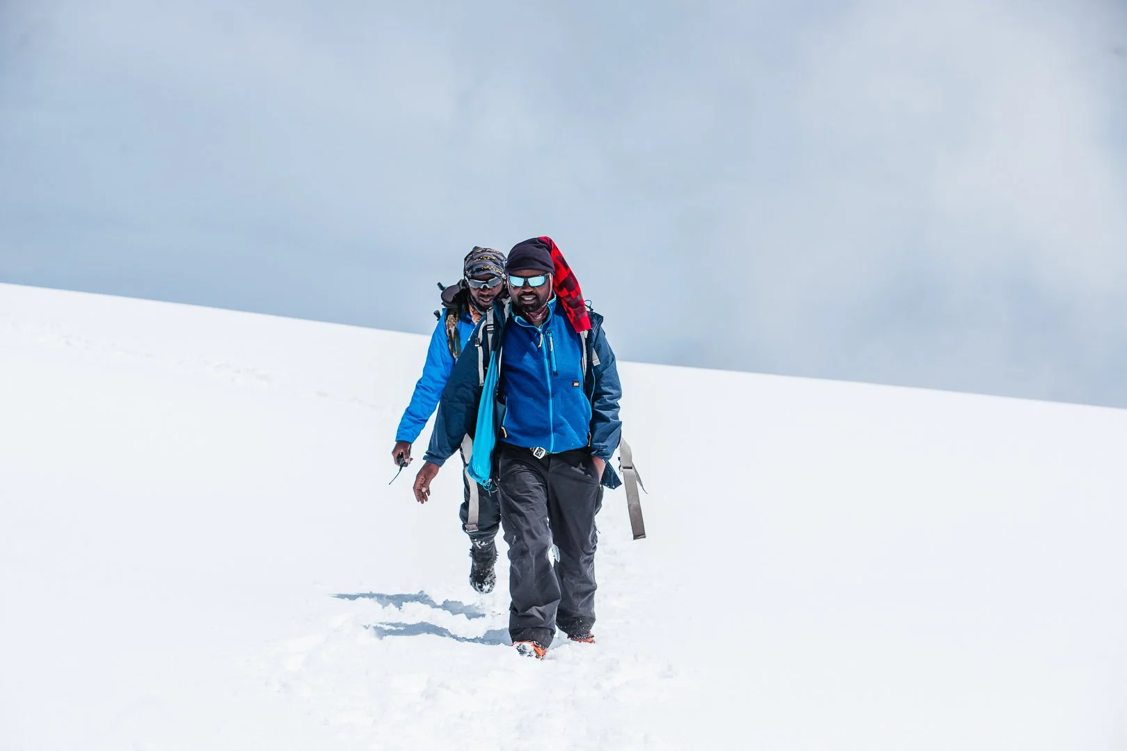 Two men hiking through snow in a snowy landscape, dressed in winter gear including jackets, hats, and sunglasses.