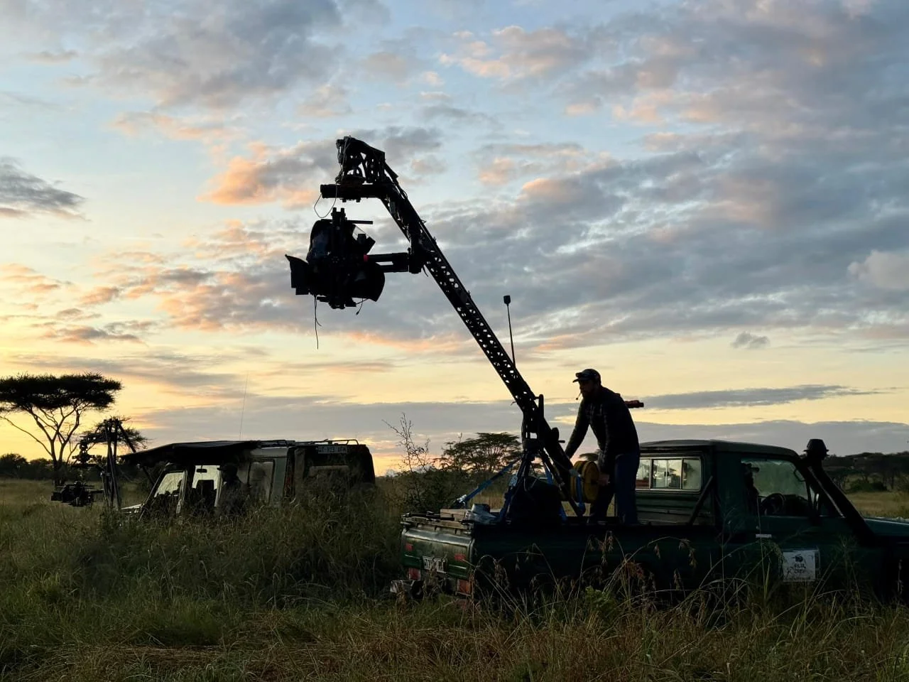 A person operating camera equipment mounted on a long crane from the back of a truck during sunset in a grassy field with sparse trees.