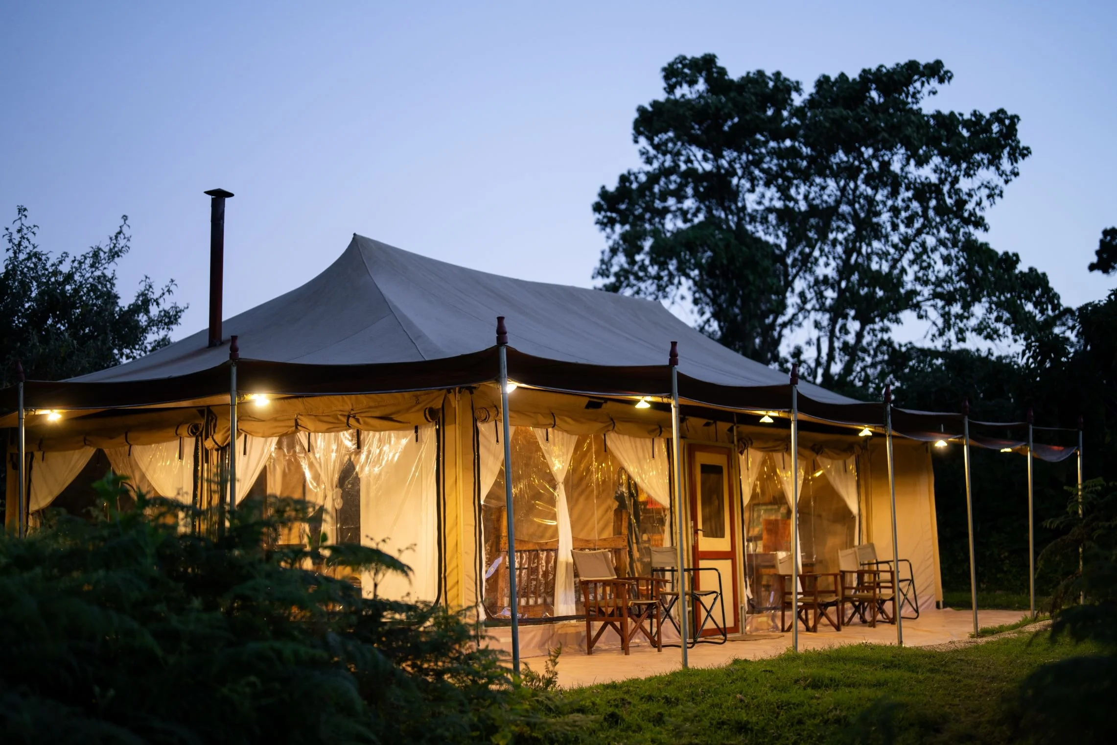 A large outdoor event tent illuminated from inside, set up in a grassy area with surrounding trees, during dusk or early evening.