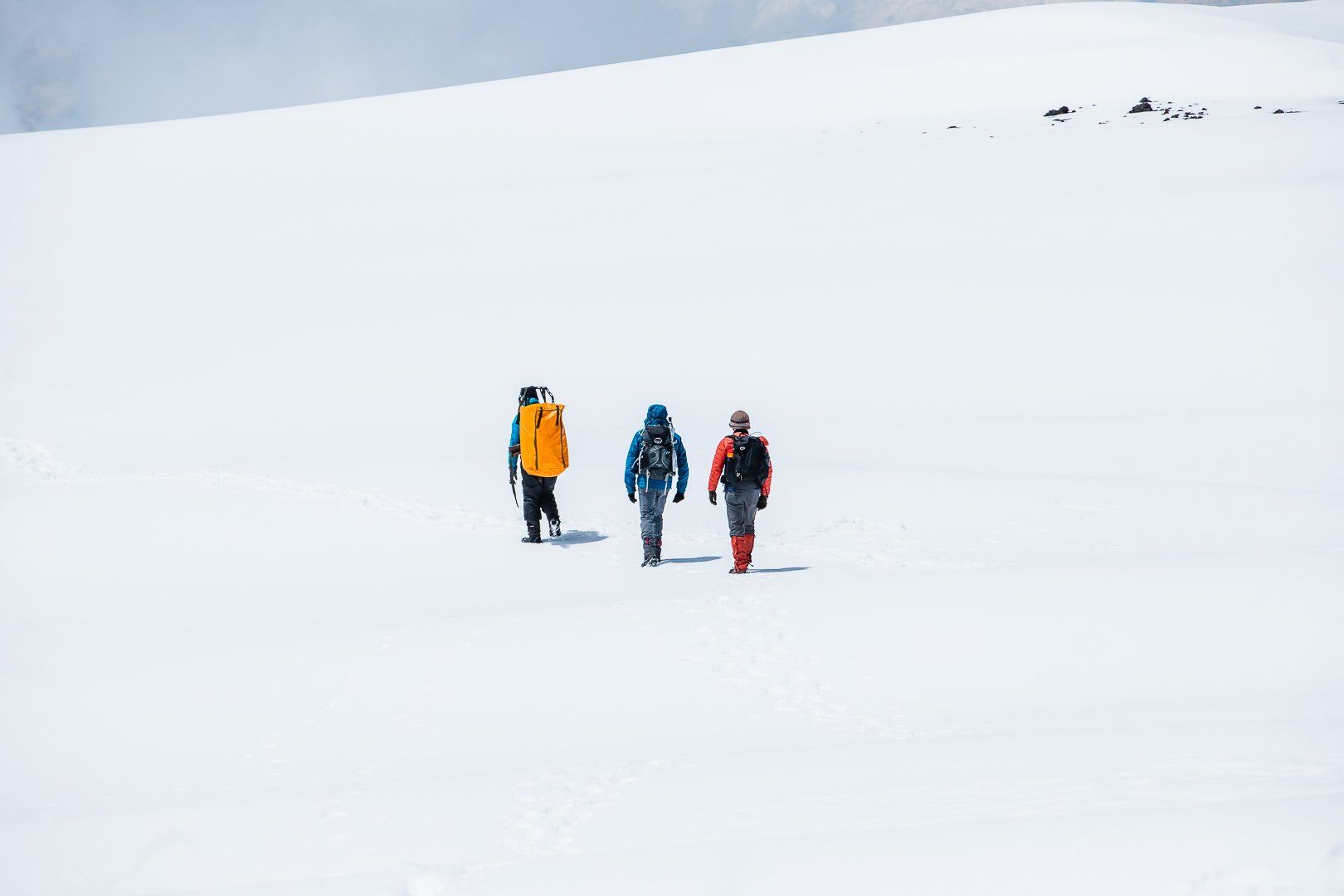 Three hikers walking through a snow-covered landscape, wearing winter gear and backpacks, with a clear sky above.