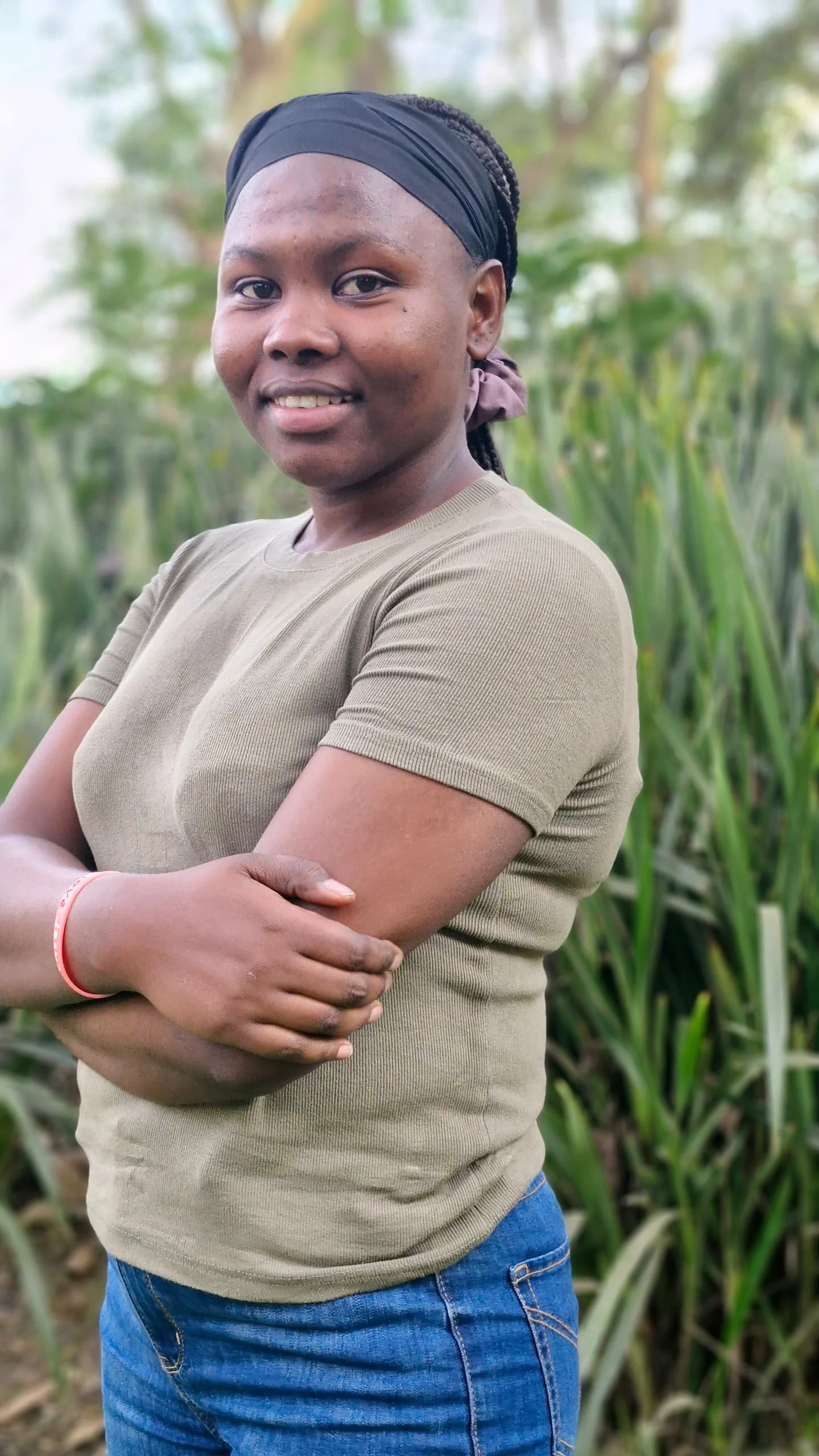 A woman standing outdoors in a field of tall green plants, smiling with her arms crossed, wearing a beige t-shirt and blue jeans, with braided hair covered by a black headscarf.