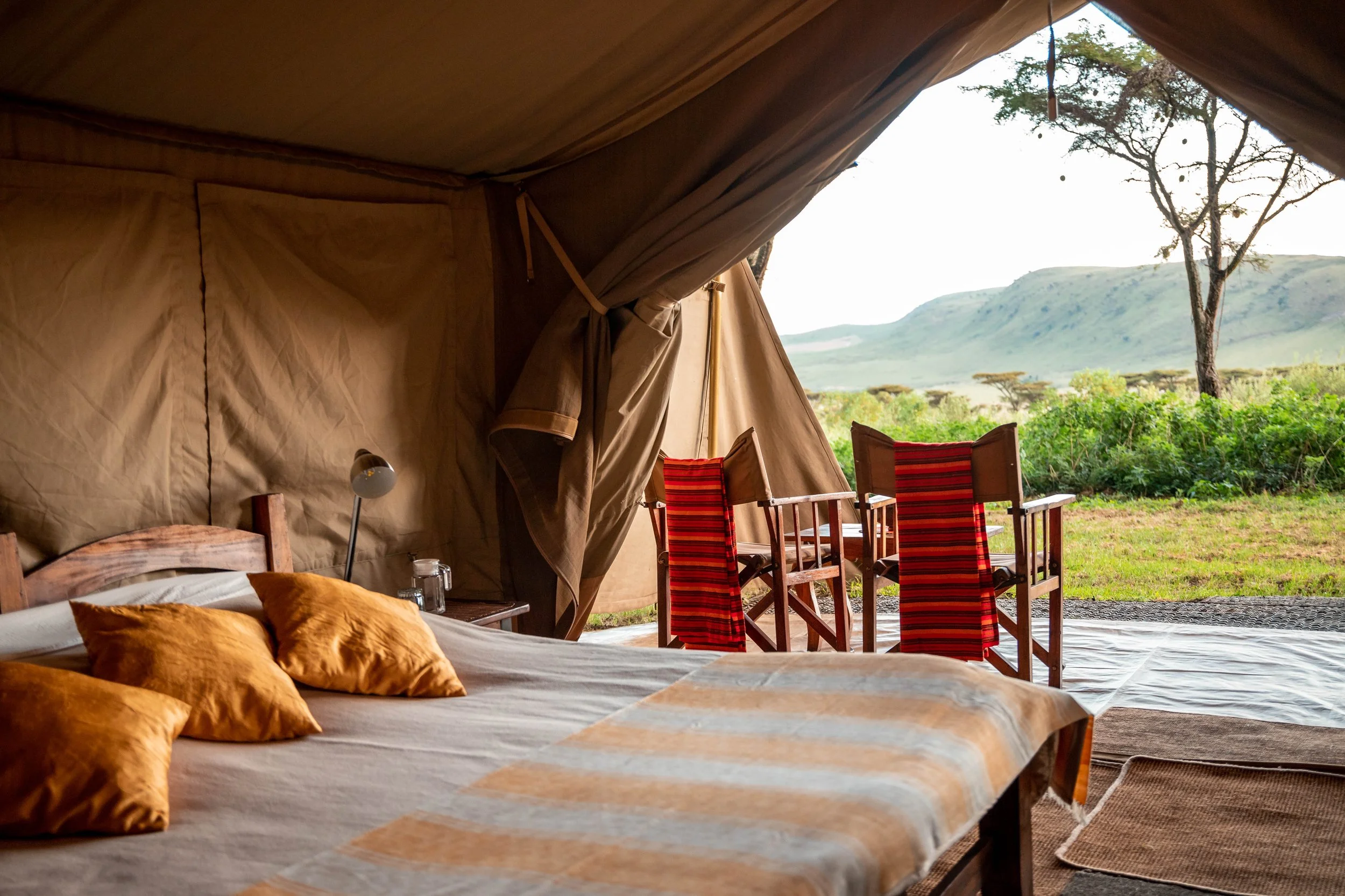 Interior view of a safari tent with beds, orange pillows, a small lamp, and a table. The tent has an open side showing outdoor landscape with trees, grass, and distant hills.