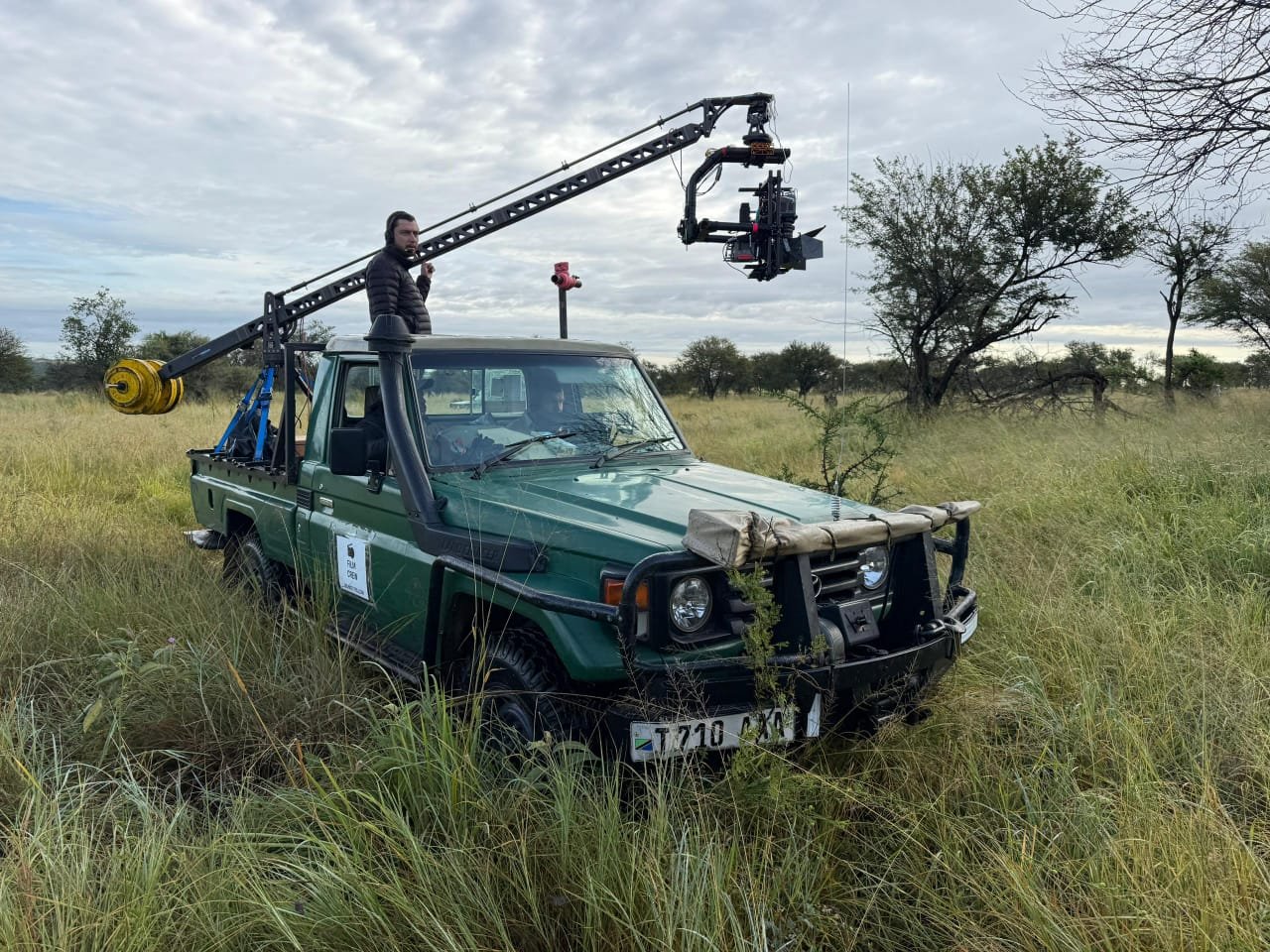 A green safari vehicle with a mounted camera crane in tall grass, in a safari landscape with trees and a cloudy sky.
