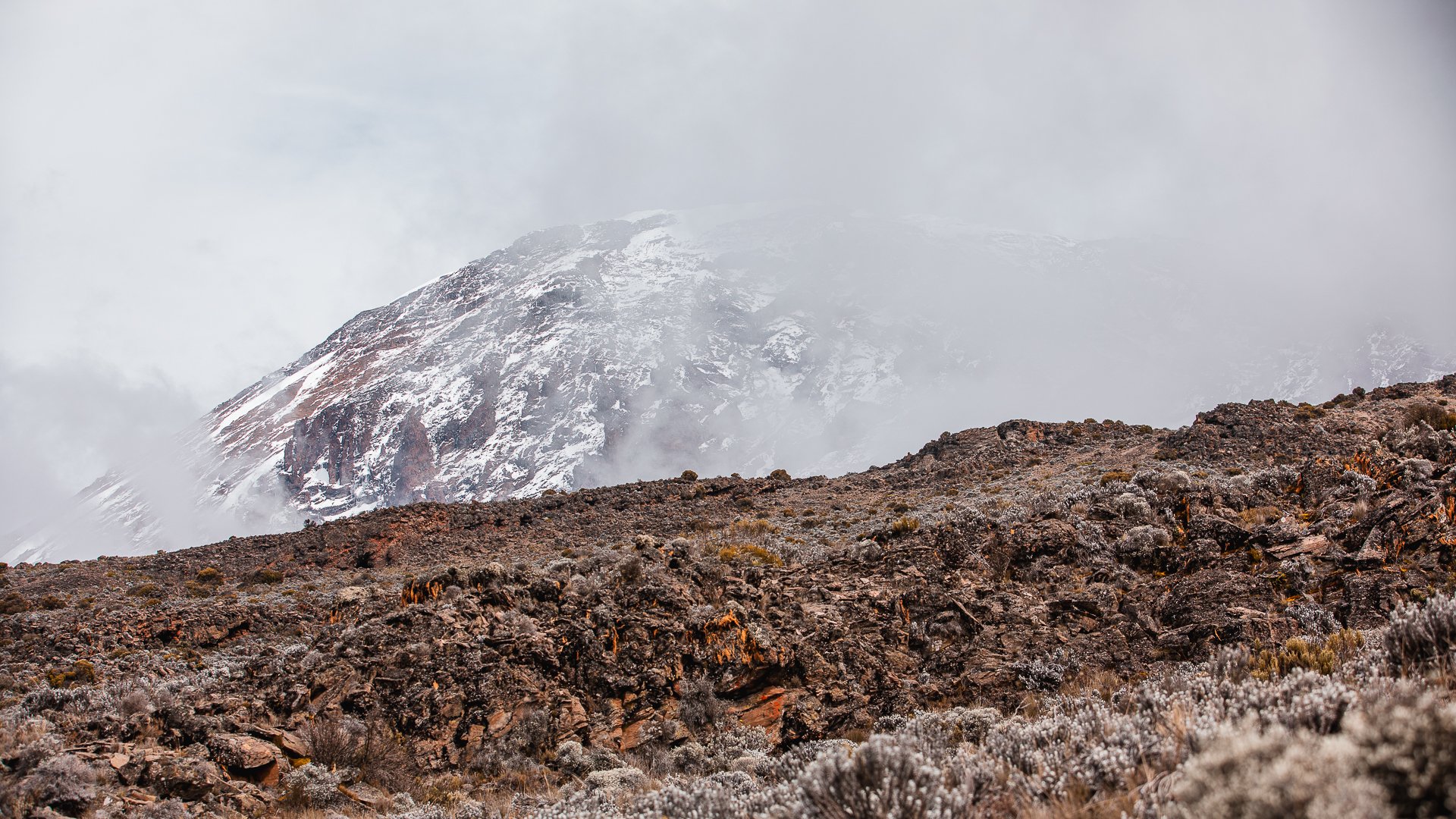 Snow-capped mountain partially obscured by clouds, with rocky terrain and sparse shrubbery in the foreground.