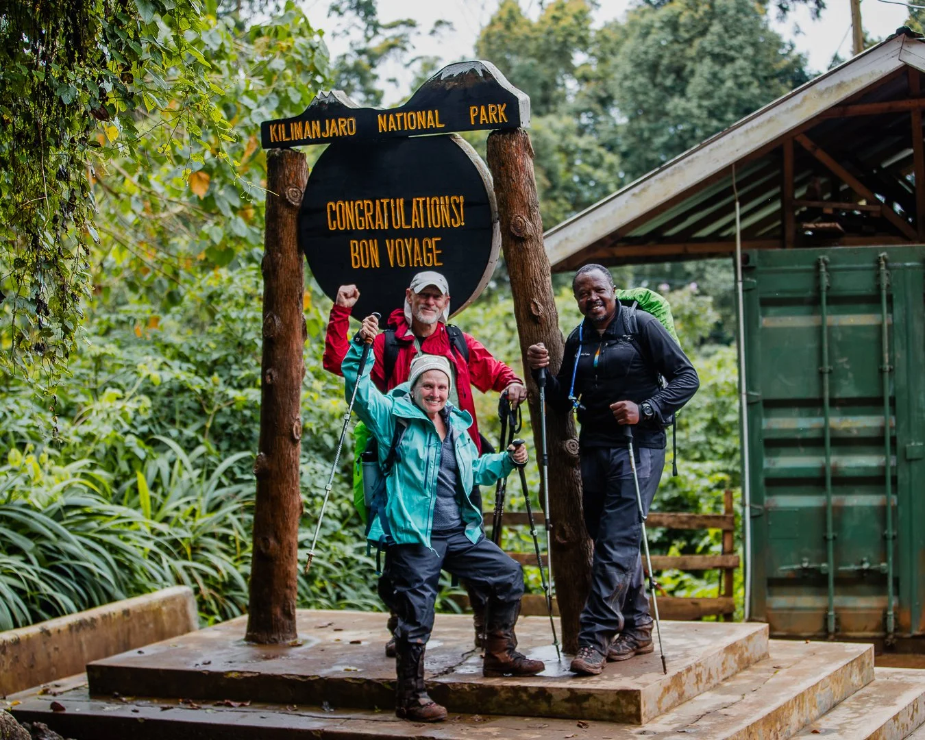 Three hikers standing on a platform at Kilimanjaro National Park, celebrating their successful climb, with a sign behind them that reads 'Congratulations! Bon Voyage' and the park's name.