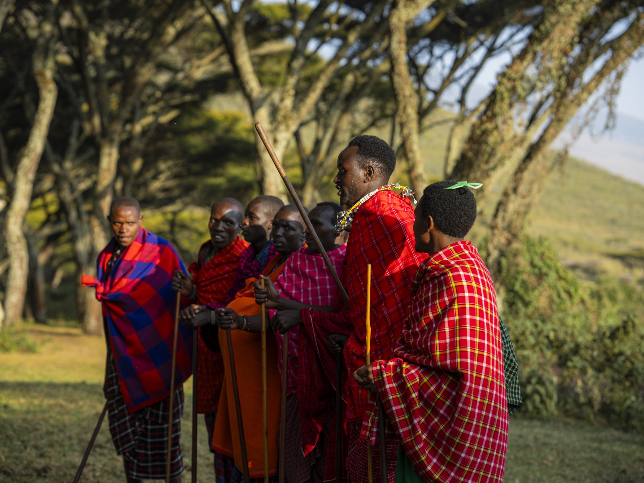 A group of indigenous people dressed in colorful traditional clothing, holding spears, standing in an outdoor setting with trees and greenery in the background.