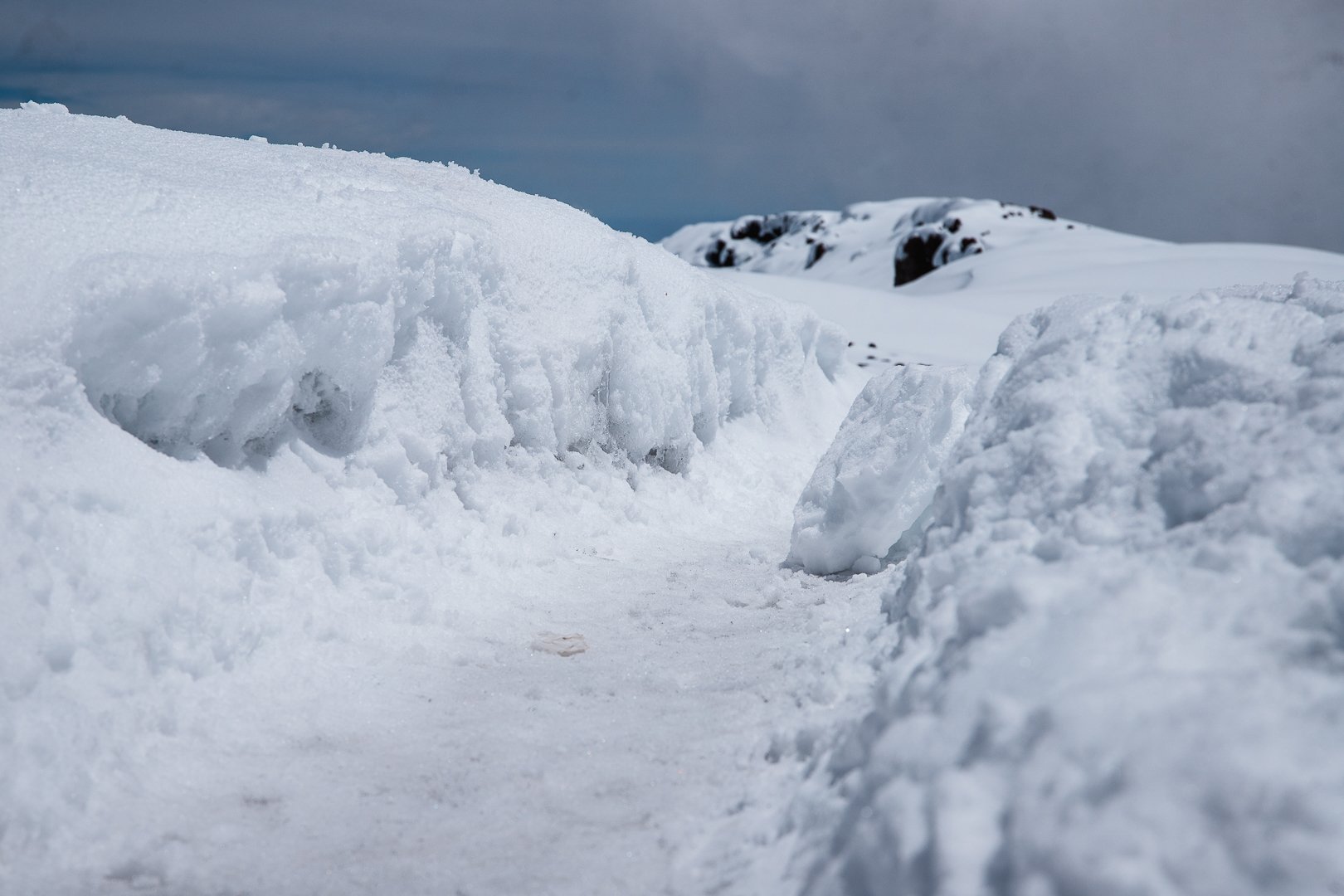 Snow-covered path flanked by high snowbanks with mountains in the background and a cloudy sky.