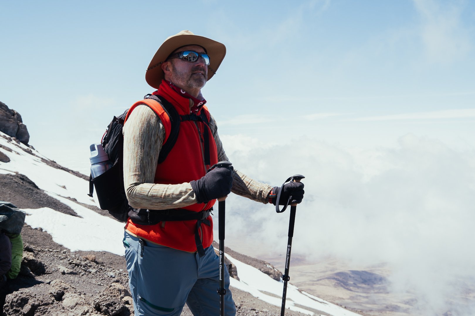 A man hiking on a snowy mountain trail with mountains in the background, wearing a hat, sunglasses, red vest, and carrying trekking poles.