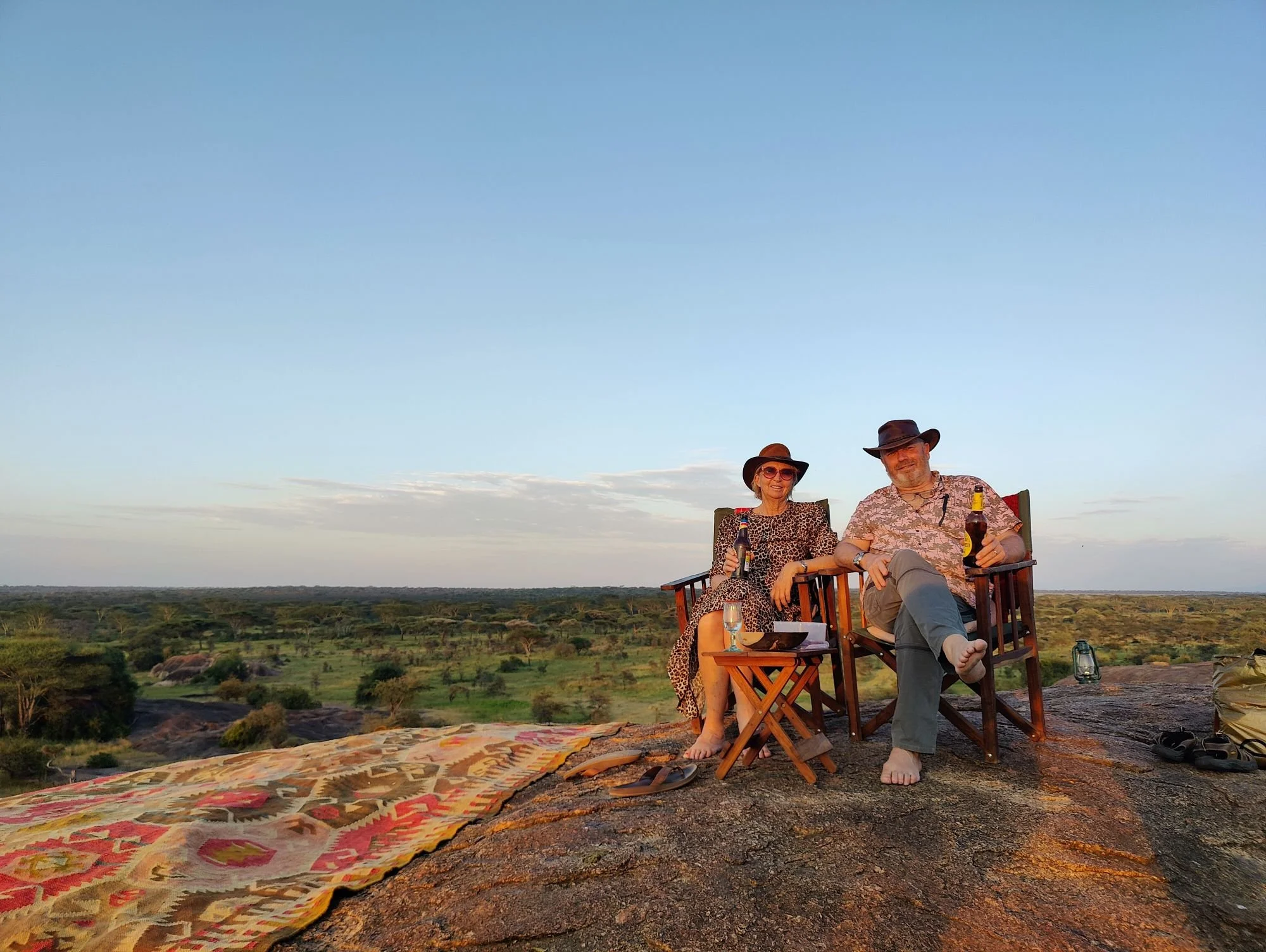 Two people, a man and a woman, sitting outdoors on a wooden bench, wearing hats and holding drinks, with a scenic landscape of trees and open land under a clear blue sky in the background.