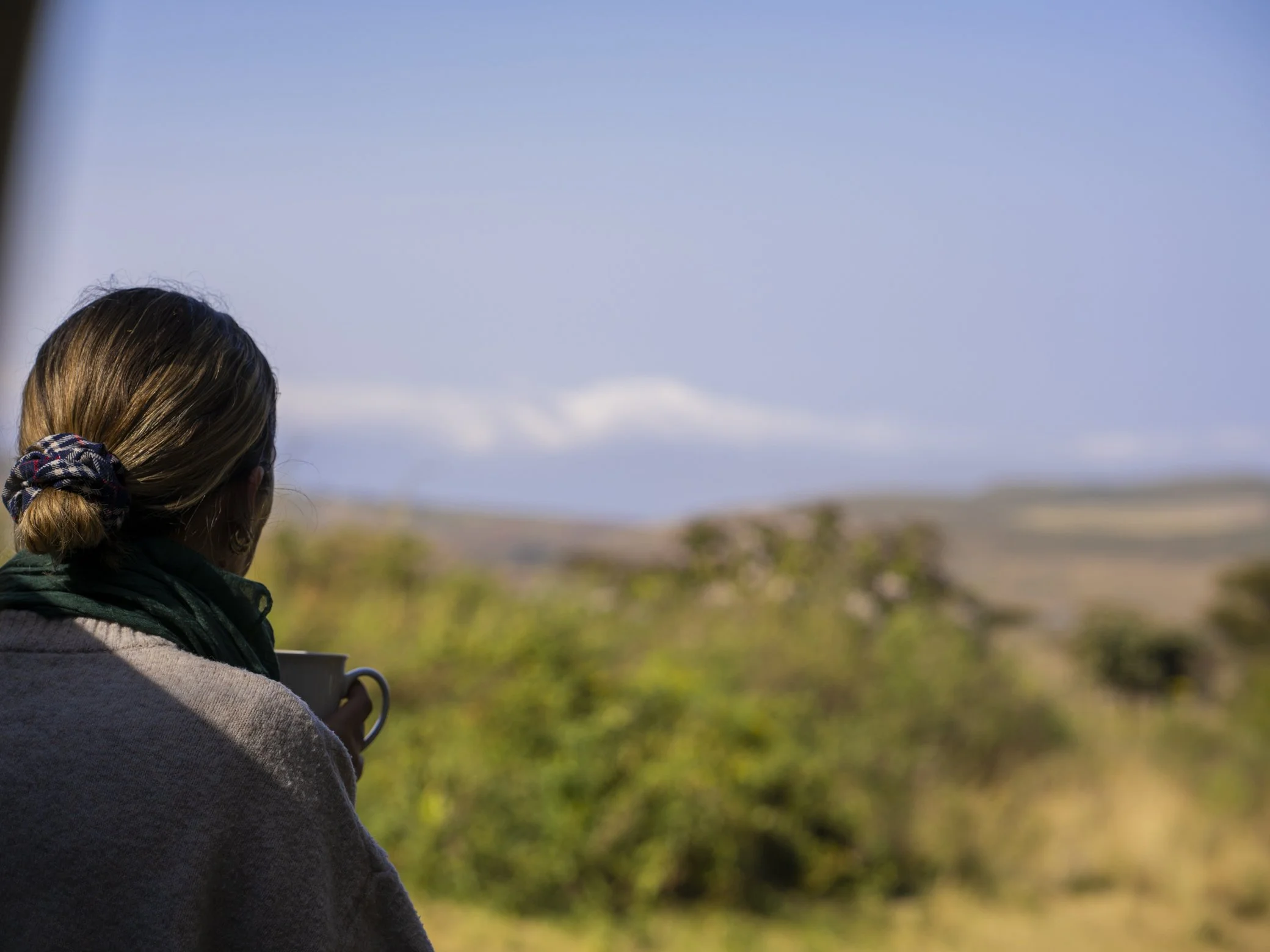 A person with long brown hair tied back with a checkered hair tie, holding a coffee mug, standing outdoors with a blurred natural landscape and a partly cloudy sky in the background.