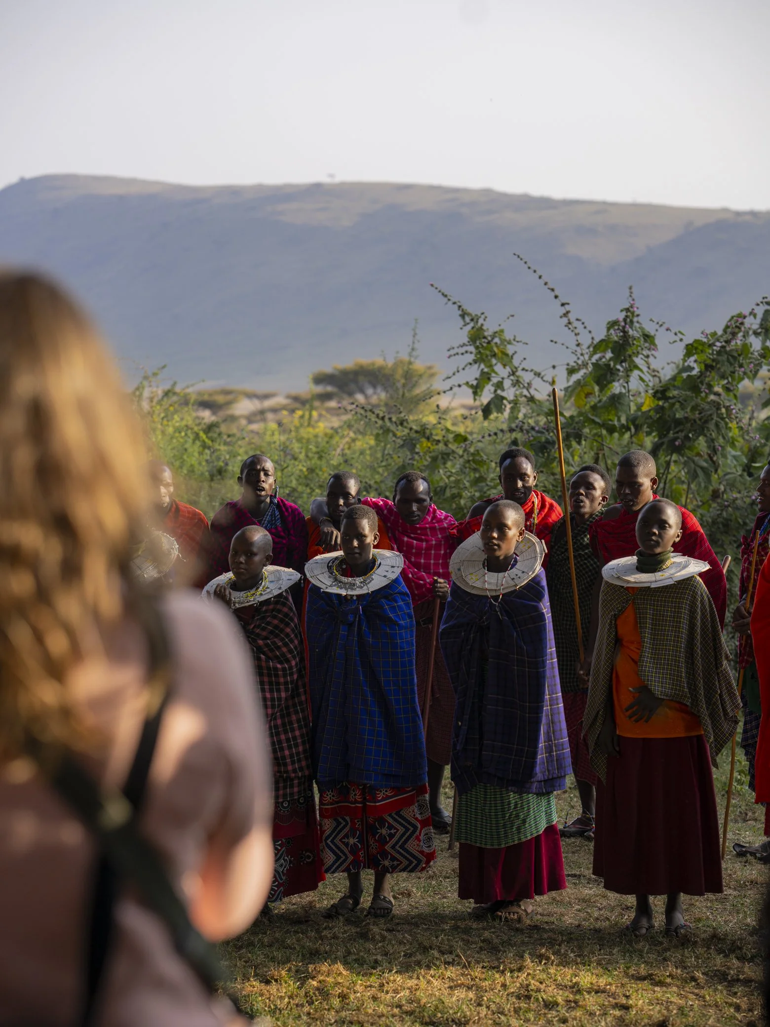 A group of Maasai people dressed in traditional clothing, standing outdoors with a hilly landscape and vegetation in the background.