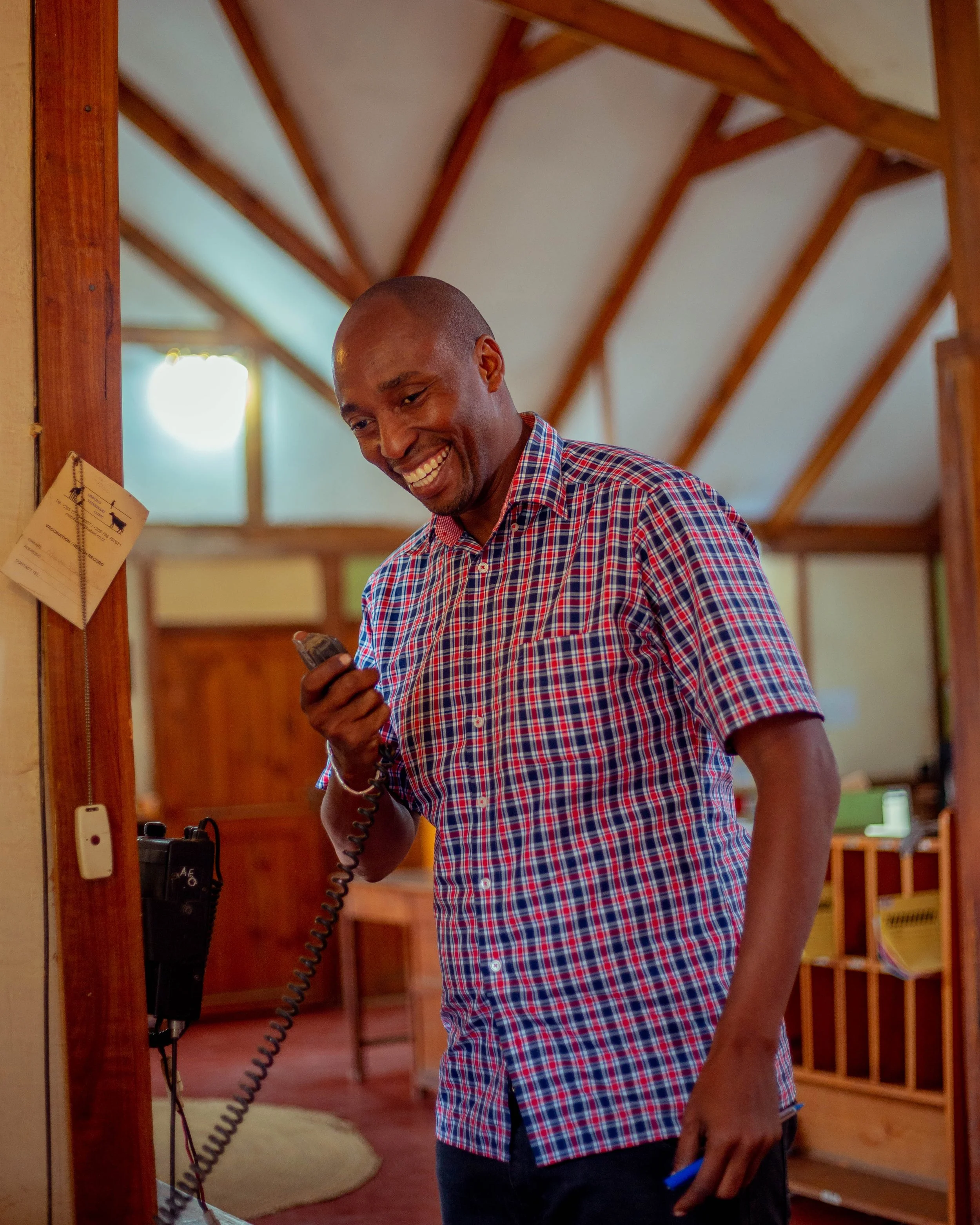 A smiling man with dark skin, wearing a red and blue checkered shirt, is holding a phone and talking on a landline phone in a room with wooden beams on the ceiling and a wooden frame around the doorway.