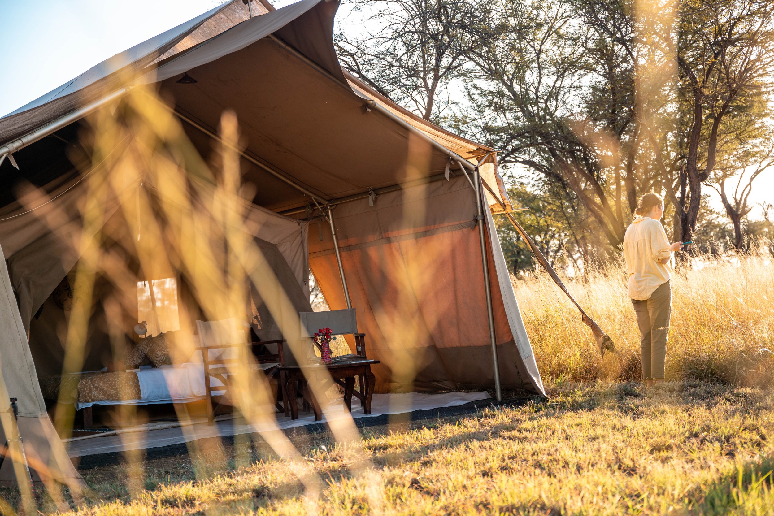 A large camping tent set up in a grassy area with a woman standing nearby, holding a phone, during what appears to be late afternoon or early evening with sunlight filtering through trees.