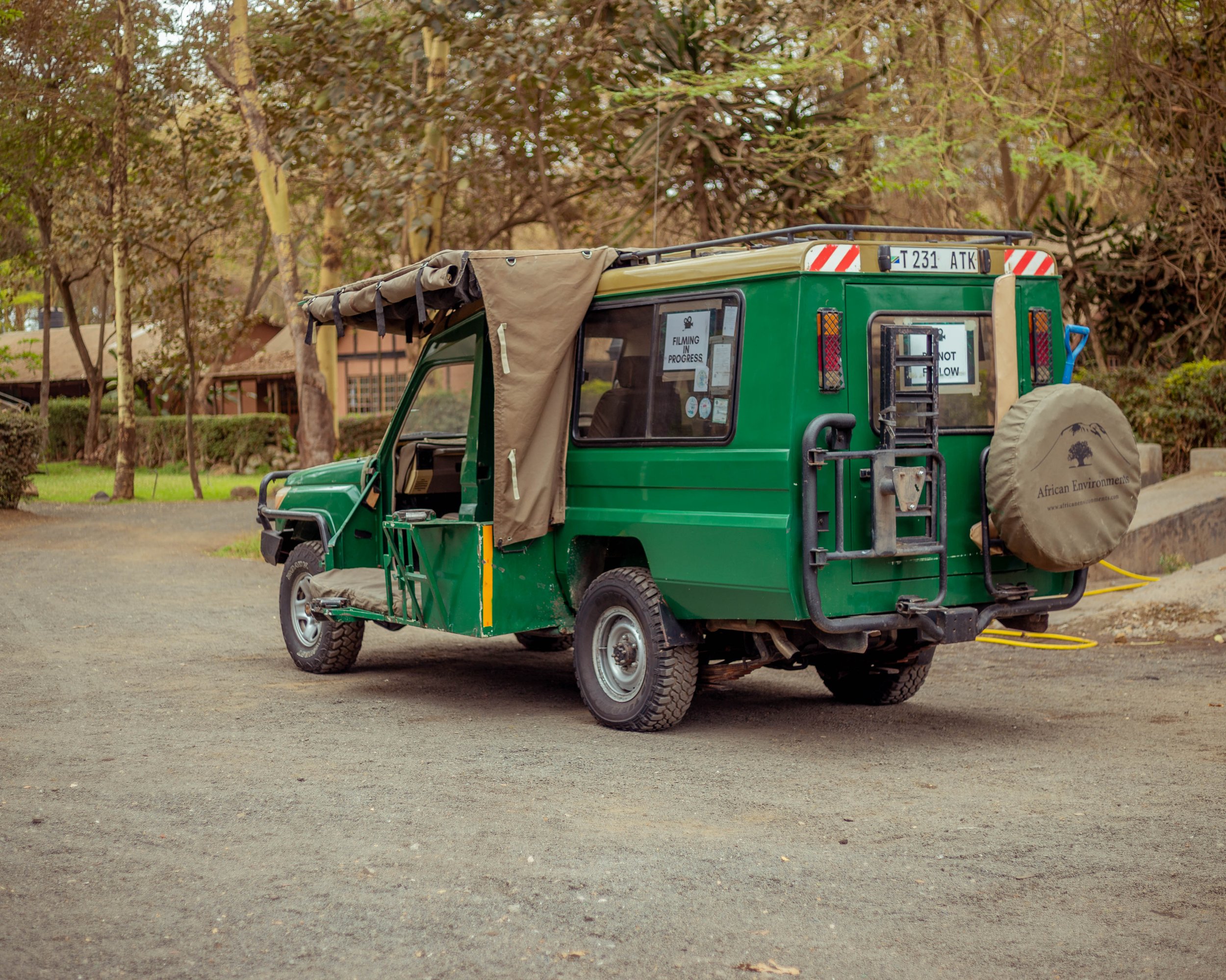 A green safari vehicle with beige roof and African Environment logo parked on a dirt path in a forested area.