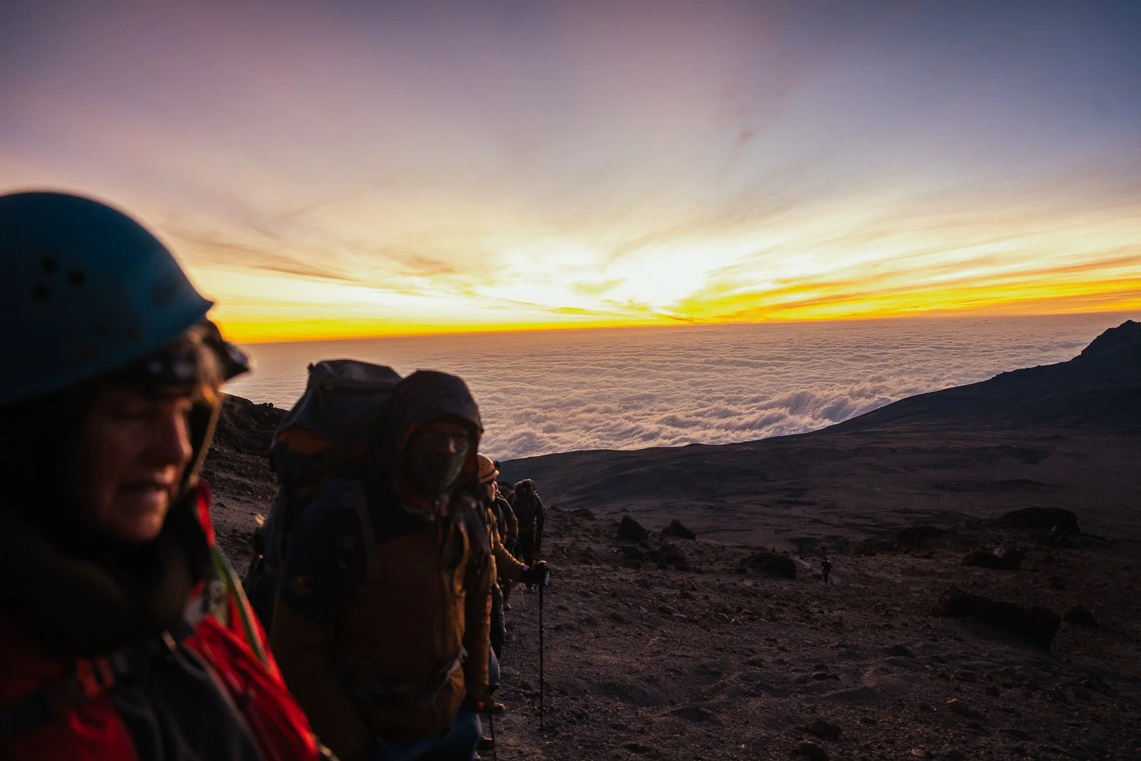 Group of hikers ascending a mountain at sunrise with a view above the clouds and a colorful sky.