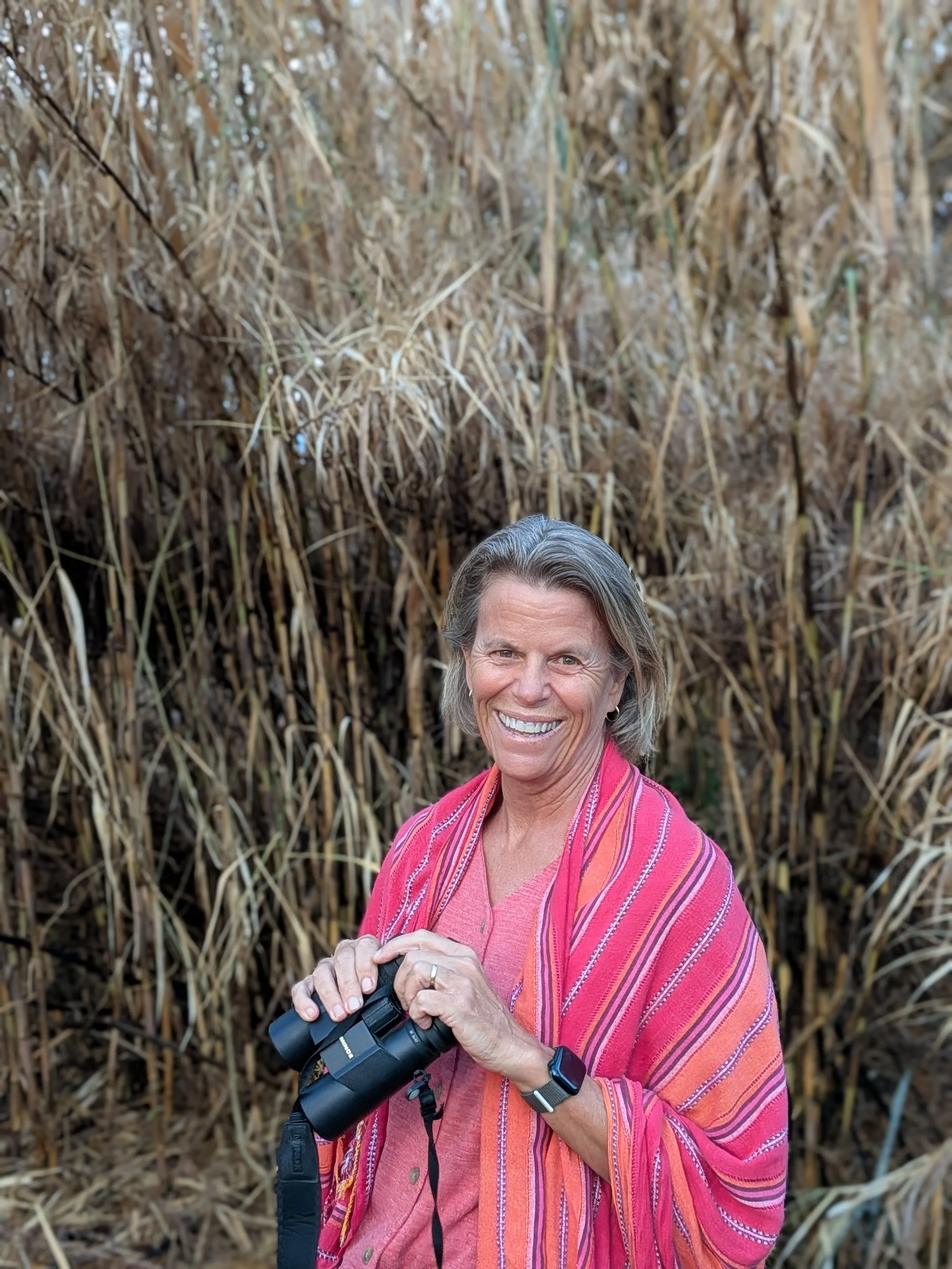 A woman with gray hair, smiling and holding binoculars, standing in front of tall dry grass or reeds, wearing a pink and orange striped shawl and a pink shirt.