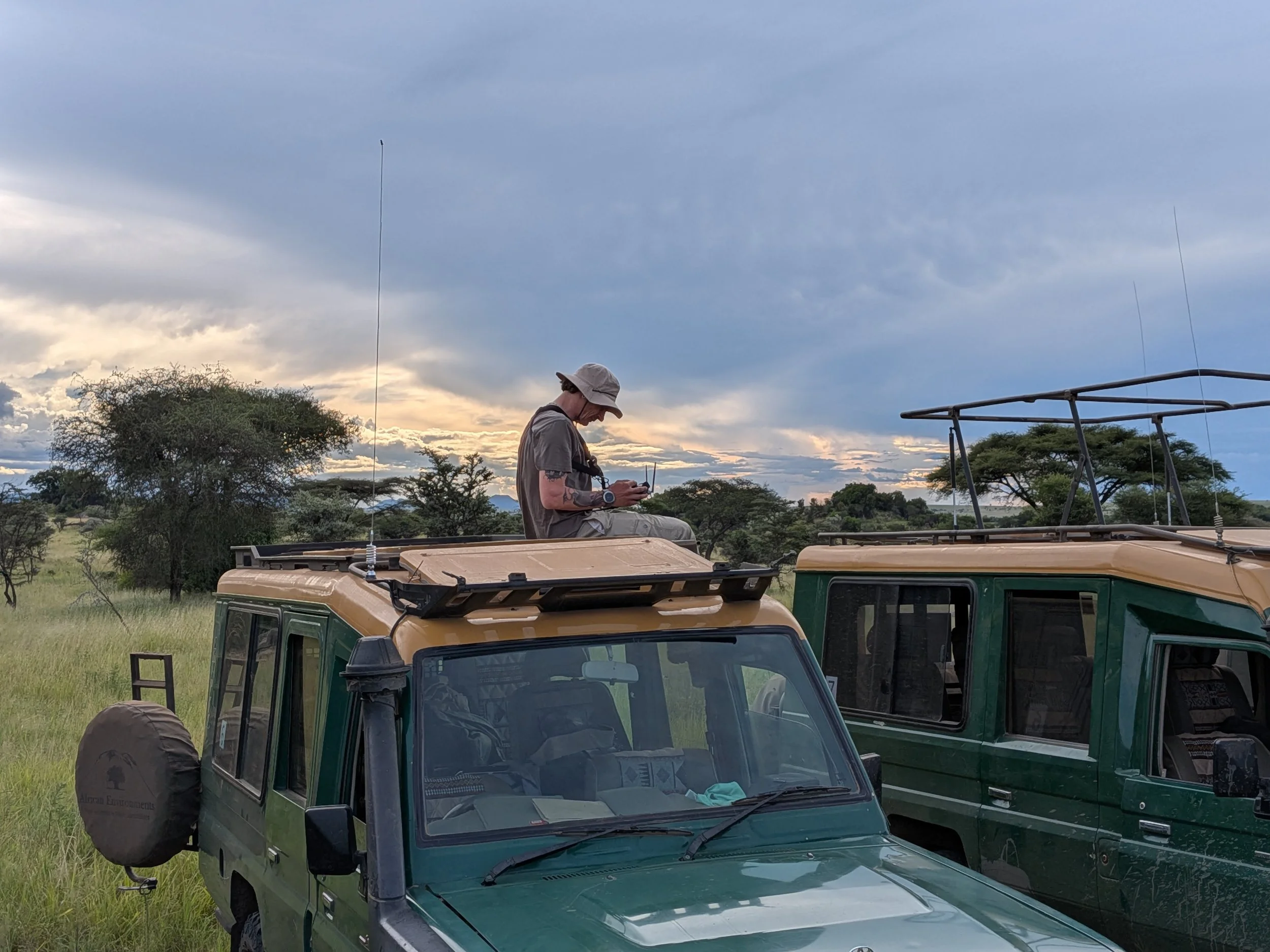 A person sitting on the roof of a safari vehicle using a remote control, with multiple safari vehicles and trees in the background, under a cloudy sky at sunset.
