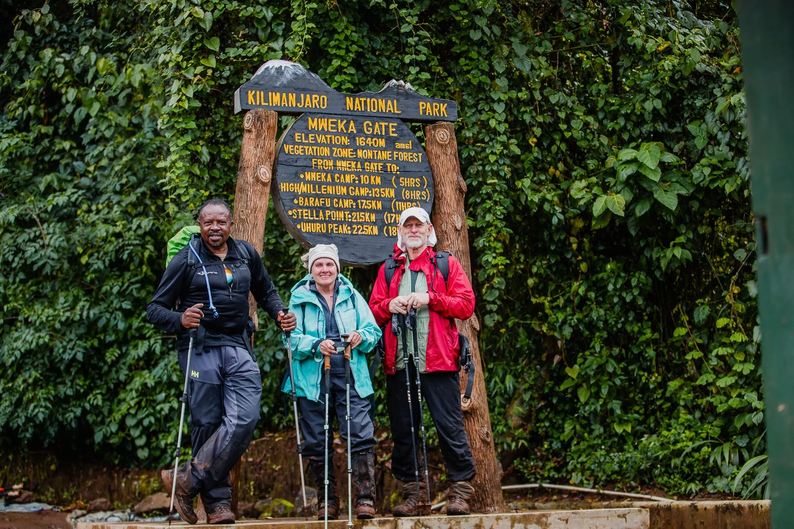 Three hikers standing in front of a wooden signboard at Kilimanjaro National Park. The sign displays elevation details and routes to various camps. The hikers are wearing outdoor gear and holding trekking poles, surrounded by lush green foliage.
