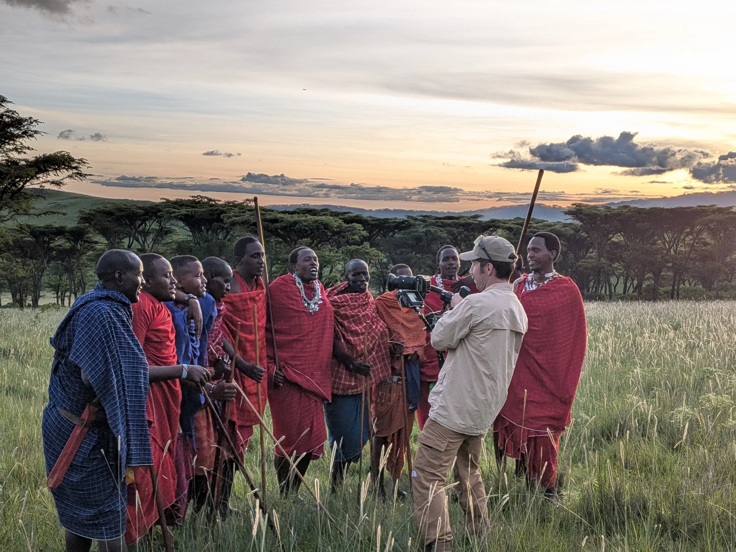 A group of Maasai men dressed in traditional red and blue clothing, with some adorned with jewelry, gather in a grassy field during sunset. A man in a beige cap and shirt is speaking to the men, with a scenic backdrop of trees and a sunset sky.