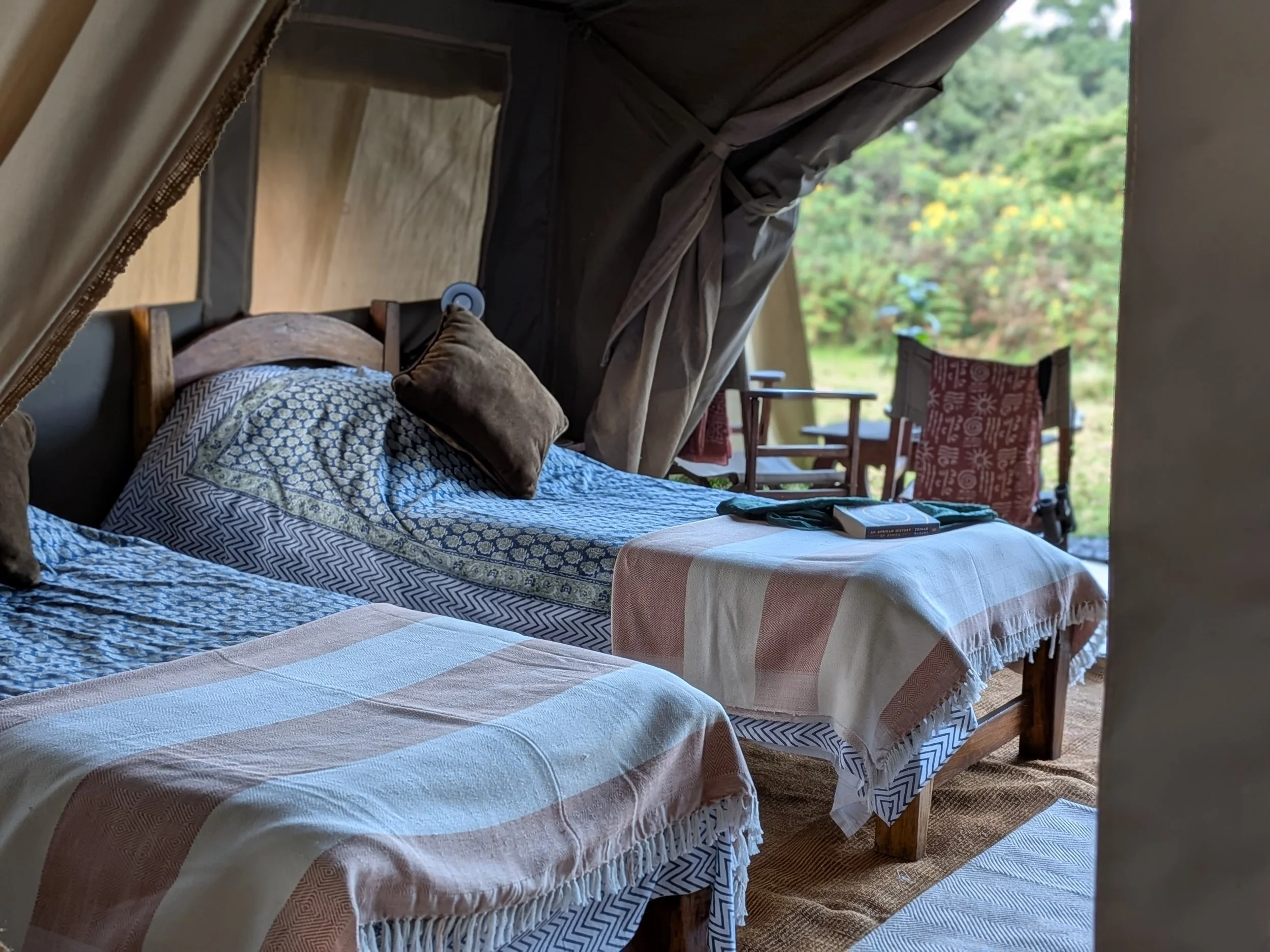 A cozy bedroom with a bed covered in patterned blue, beige, and white bedding, topped with brown and tan pillows. A small bedside lamp is attached to the head of the bed, and outside, a porch with chairs and a grassy yard with trees are visible through an open window or doorway.