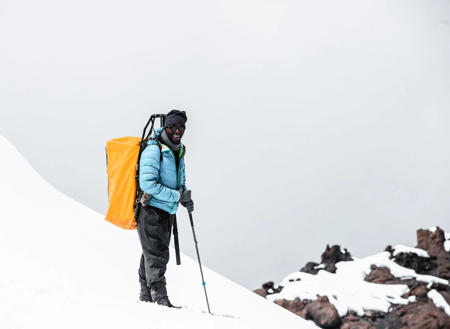 Man in winter gear standing on snowy mountain slope with snow and rocks, holding trekking poles, smiling, wearing sunglasses, a beanie, a blue jacket, and carrying an orange backpack.