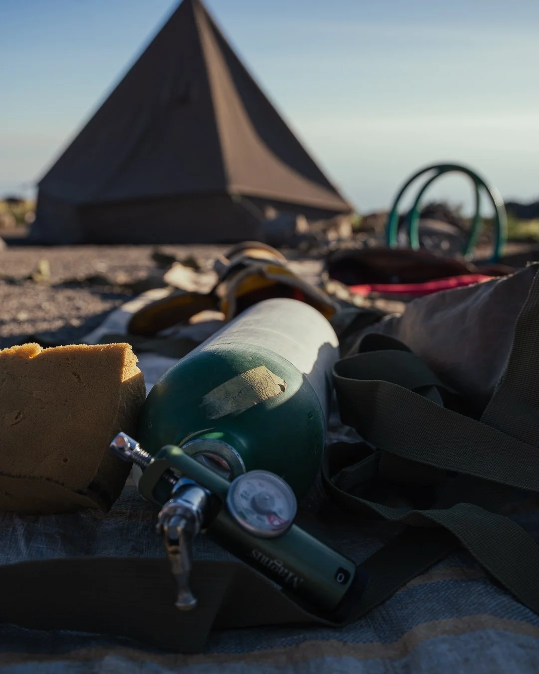 Camping scene with a tent in the background, and close-up of camping gear including a water bottle, sponge, and gauge in the foreground on the ground.