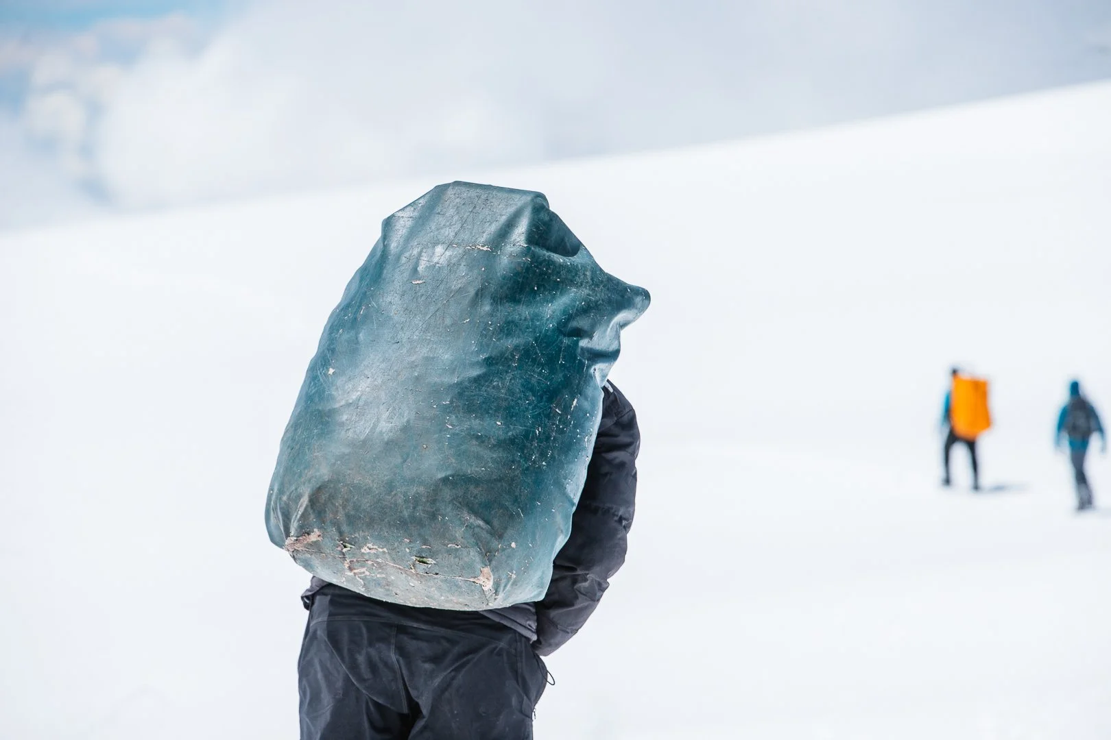 Person in black winter clothing with a large, irregularly shaped ice pack on their back, standing in a snowy landscape with other people in the distance.